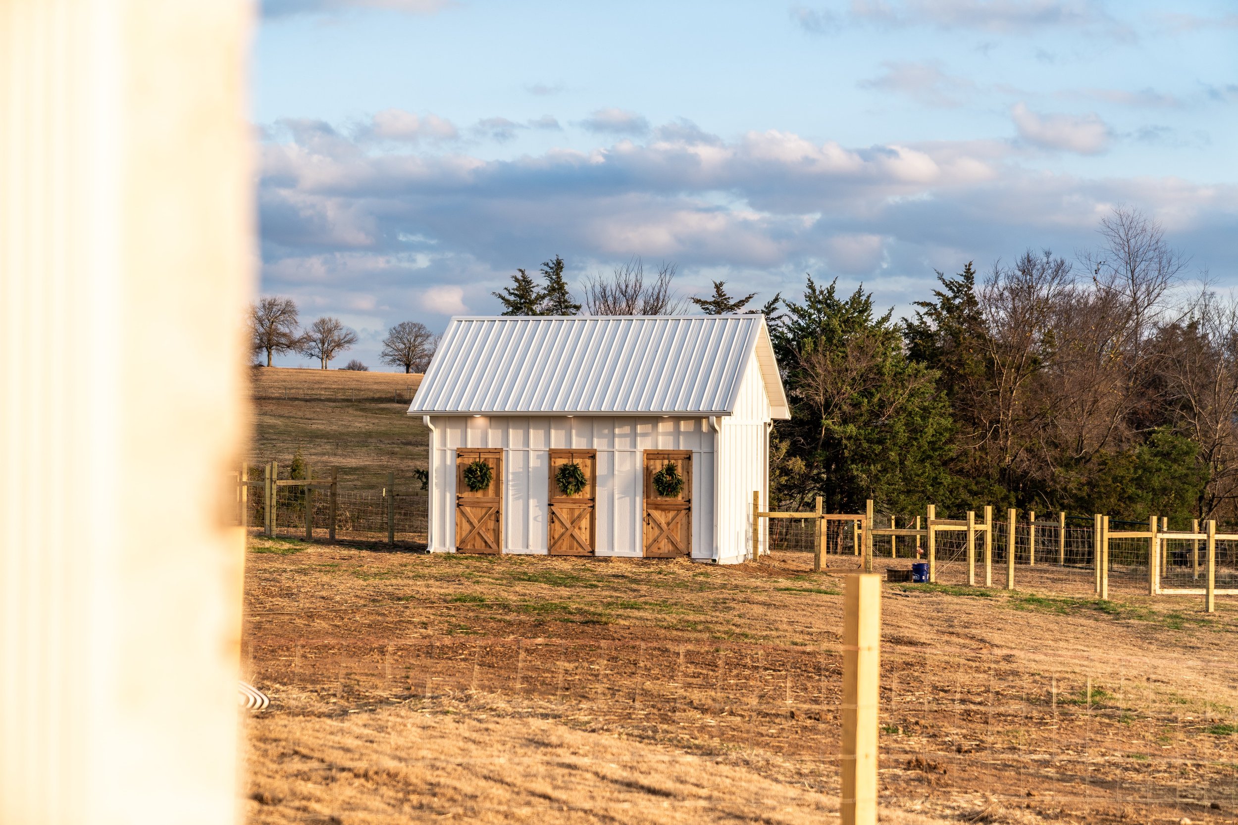 A rural scene with a small white shed decorated with three wreaths, surrounded by a wooden fence, on a sunny day with a partly cloudy sky and trees in the background.