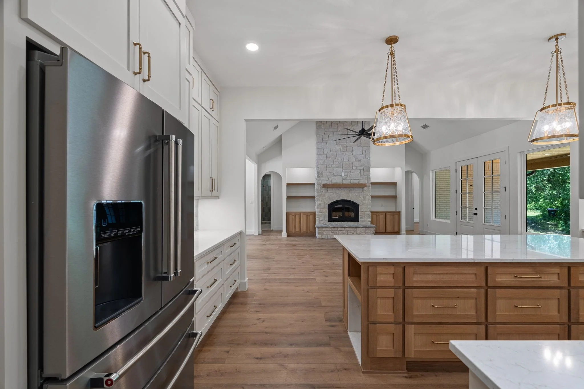 Open-concept kitchen with white cabinets, stainless steel refrigerator, kitchen island with wooden drawers, and a stone fireplace in the living area with large windows and doors leading outside.