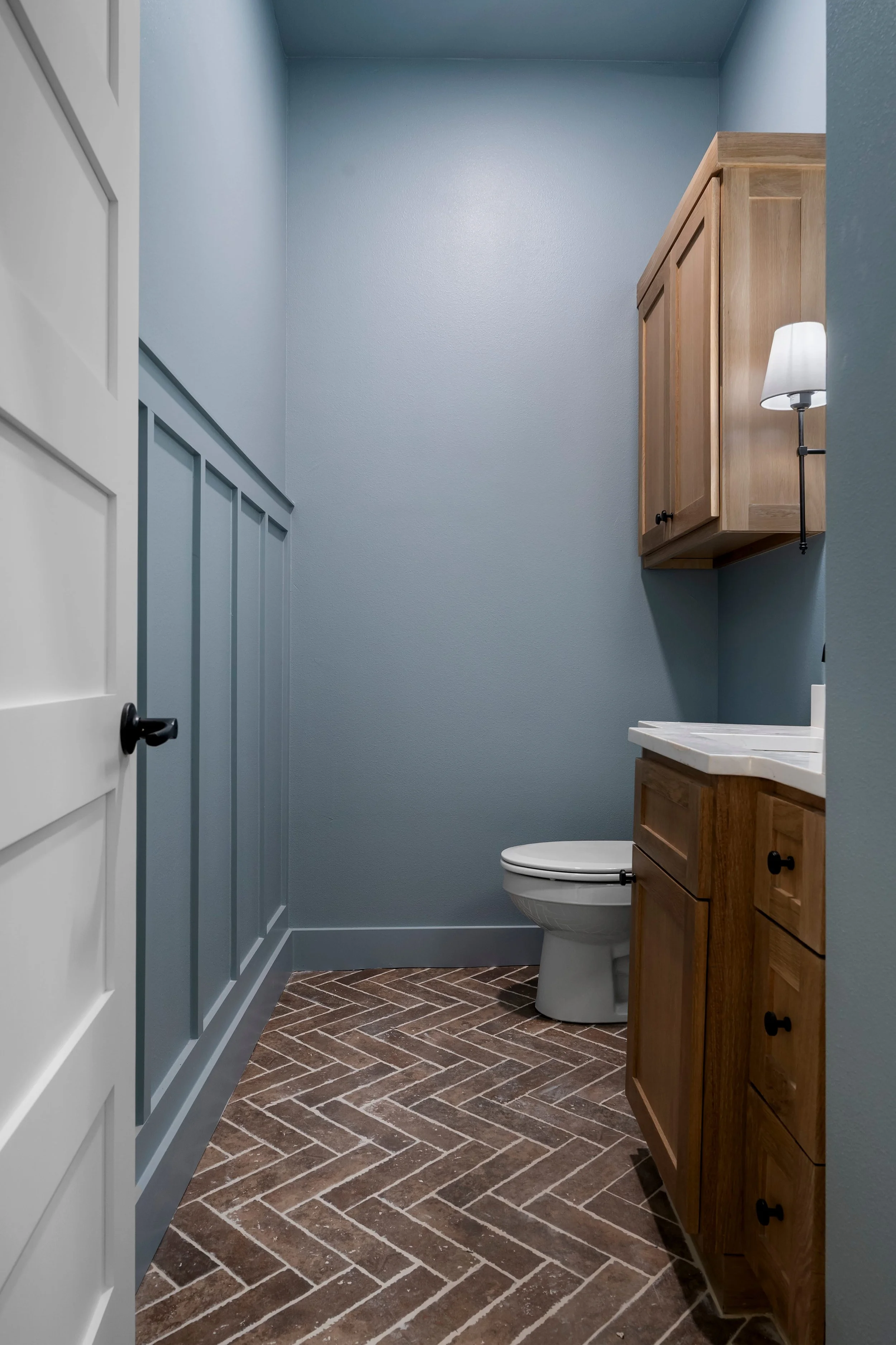 A small bathroom with blue walls, a white door, a toilet, a wooden cabinet above the toilet, and a wooden vanity with a white marble countertop. There's a wall-mounted lamp beside the cabinet and wood-look herringbone tile flooring.