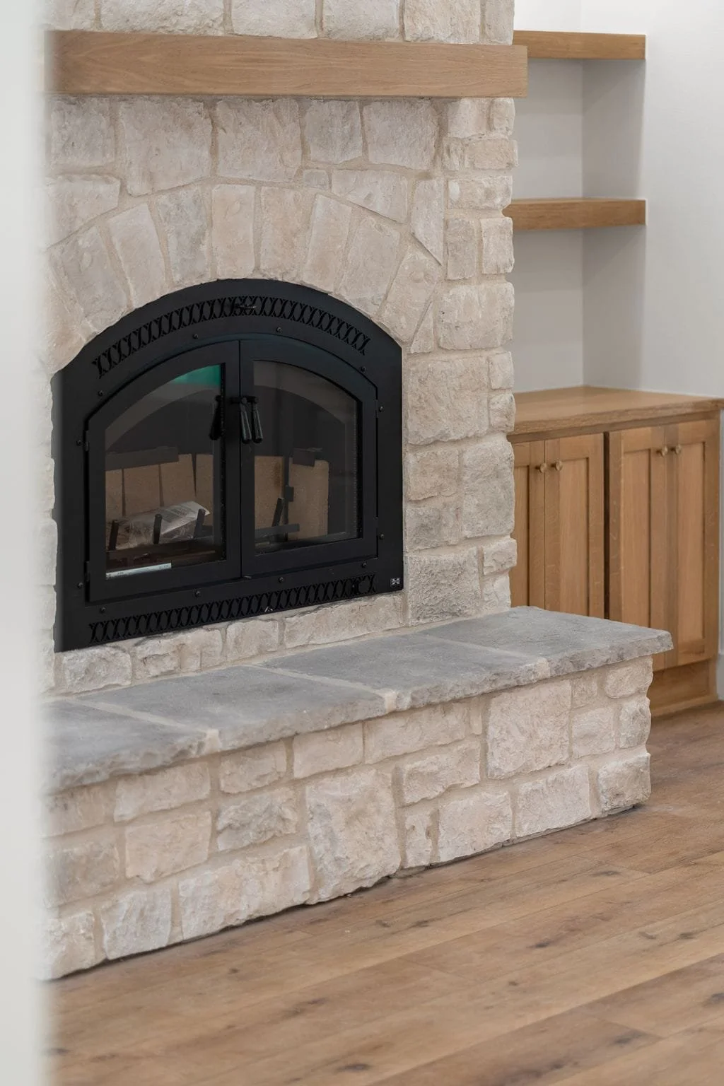 A fireplace with black metal door and stone surround in a room with hardwood floors and wooden shelves and cabinet.