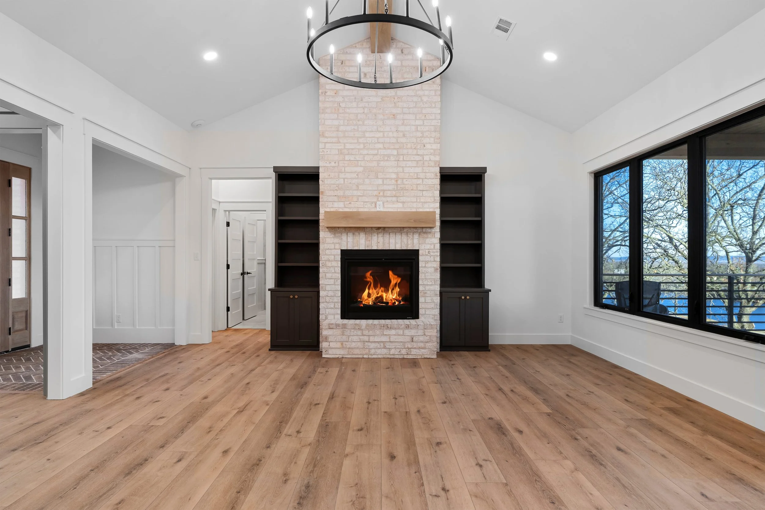 Living room with a white brick fireplace flanked by black built-in shelves, a circular chandelier above, wooden flooring, large window showing trees outside, and white walls.