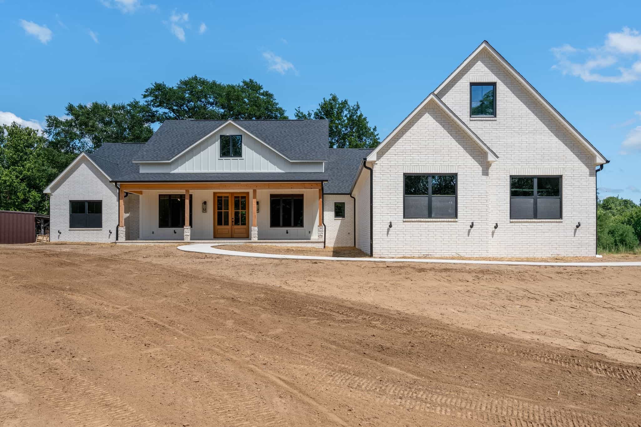 New white brick house with black window frames, wooden front door, small front porch, surrounded by a bare dirt yard, green trees in the background, under a blue sky with some clouds.