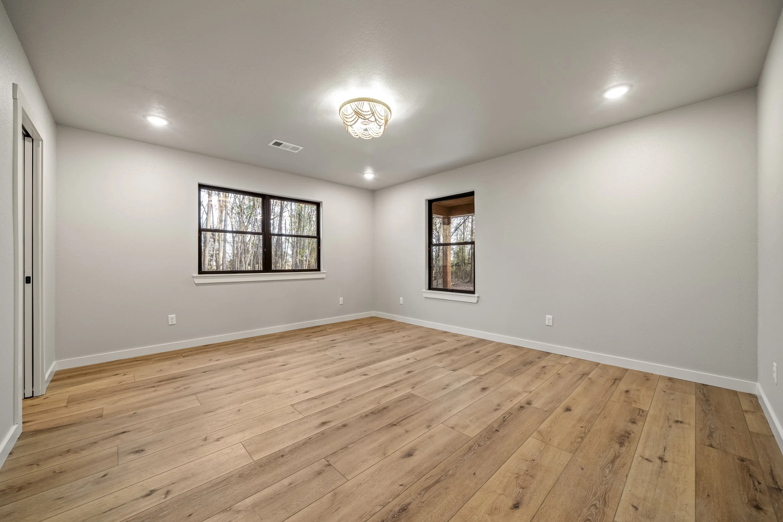 Empty room with wooden flooring, white walls, two large windows showing trees outside, ceiling lights, and a ceiling fixture.