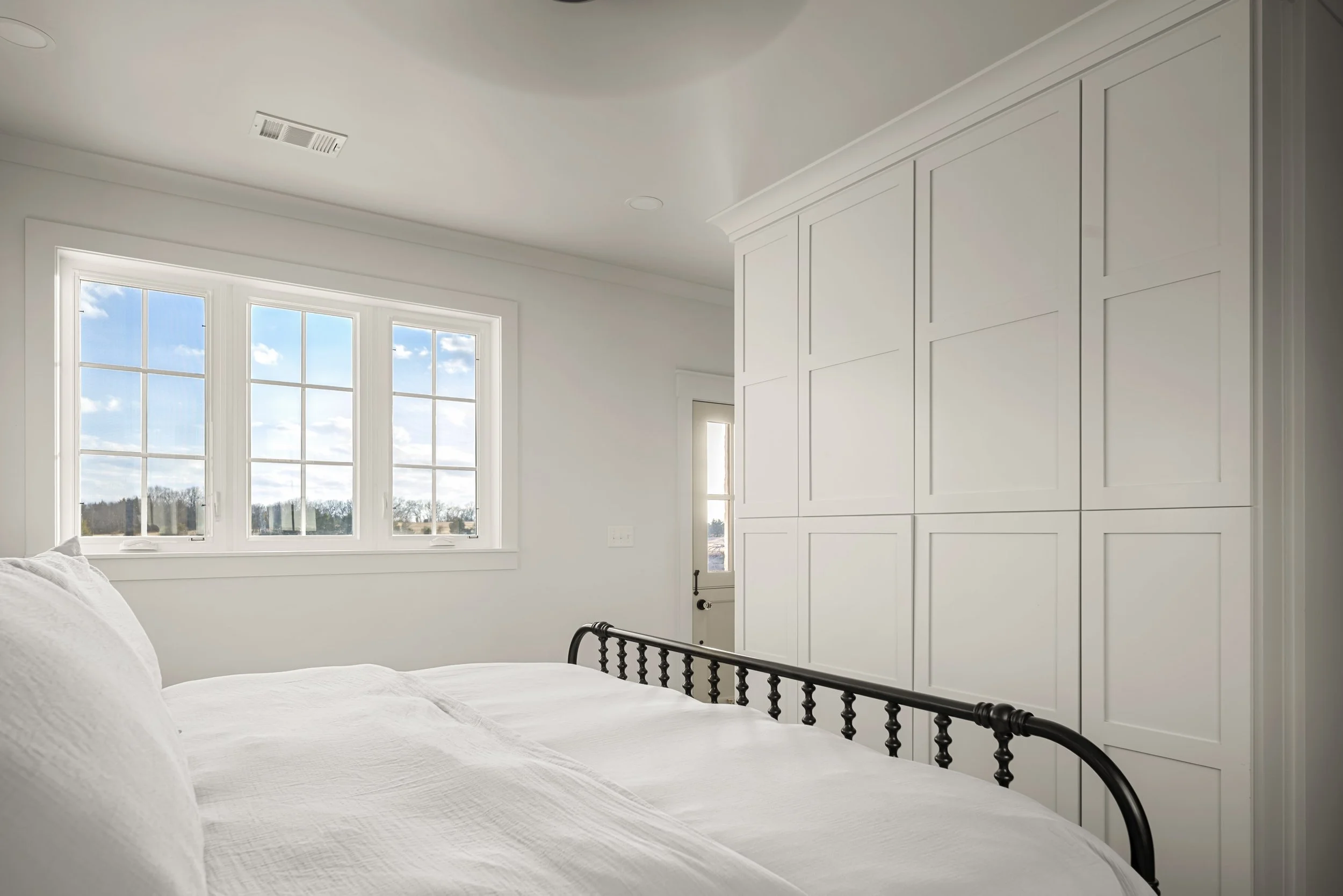 A bedroom with white walls, white bedding, a black bed frame, and a large window showing a partly cloudy sky outside.