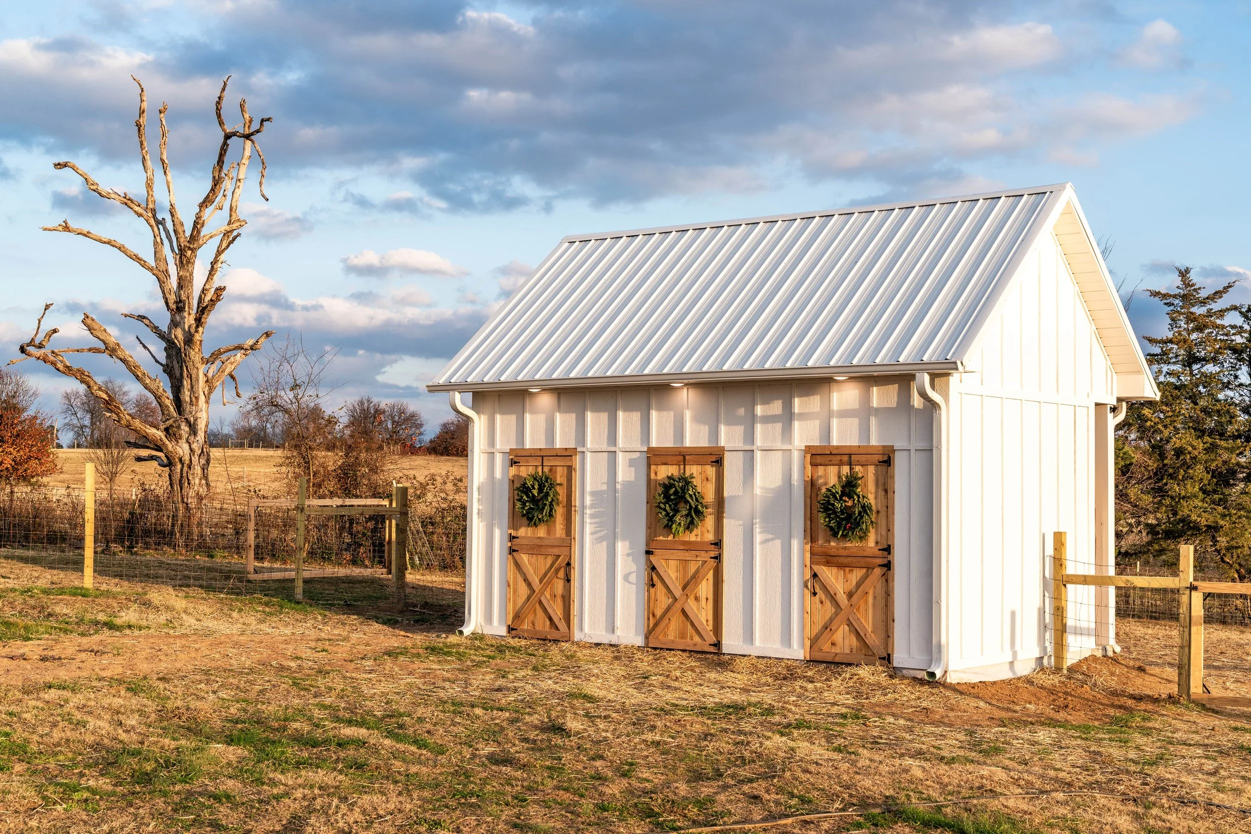 A small white shed with a metal roof, decorated with three holiday wreaths on wooden doors, standing in a rural landscape with grass, a leafless tree, and a partly cloudy sky.