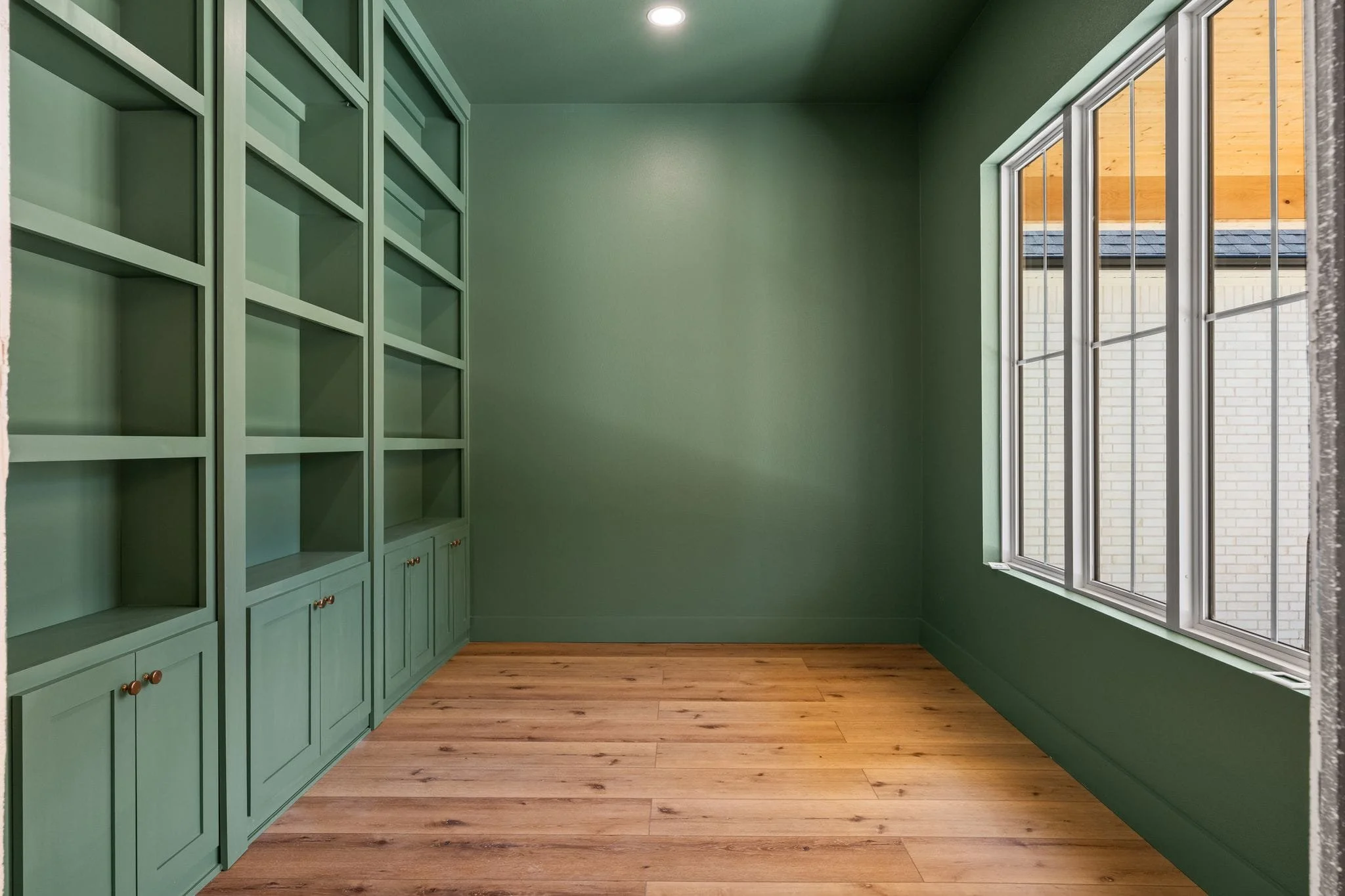 An empty room with green walls, wooden flooring, a large window with blinds, and built-in green shelving.