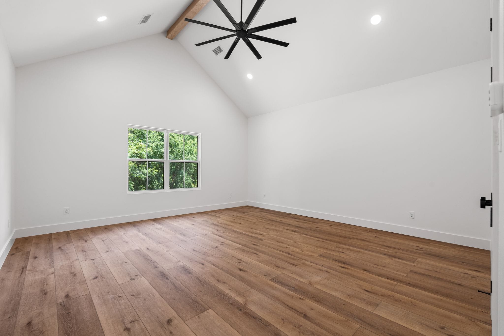 Empty room with white walls, a window showing green trees outside, hardwood flooring, a black ceiling fan, and recessed lighting.