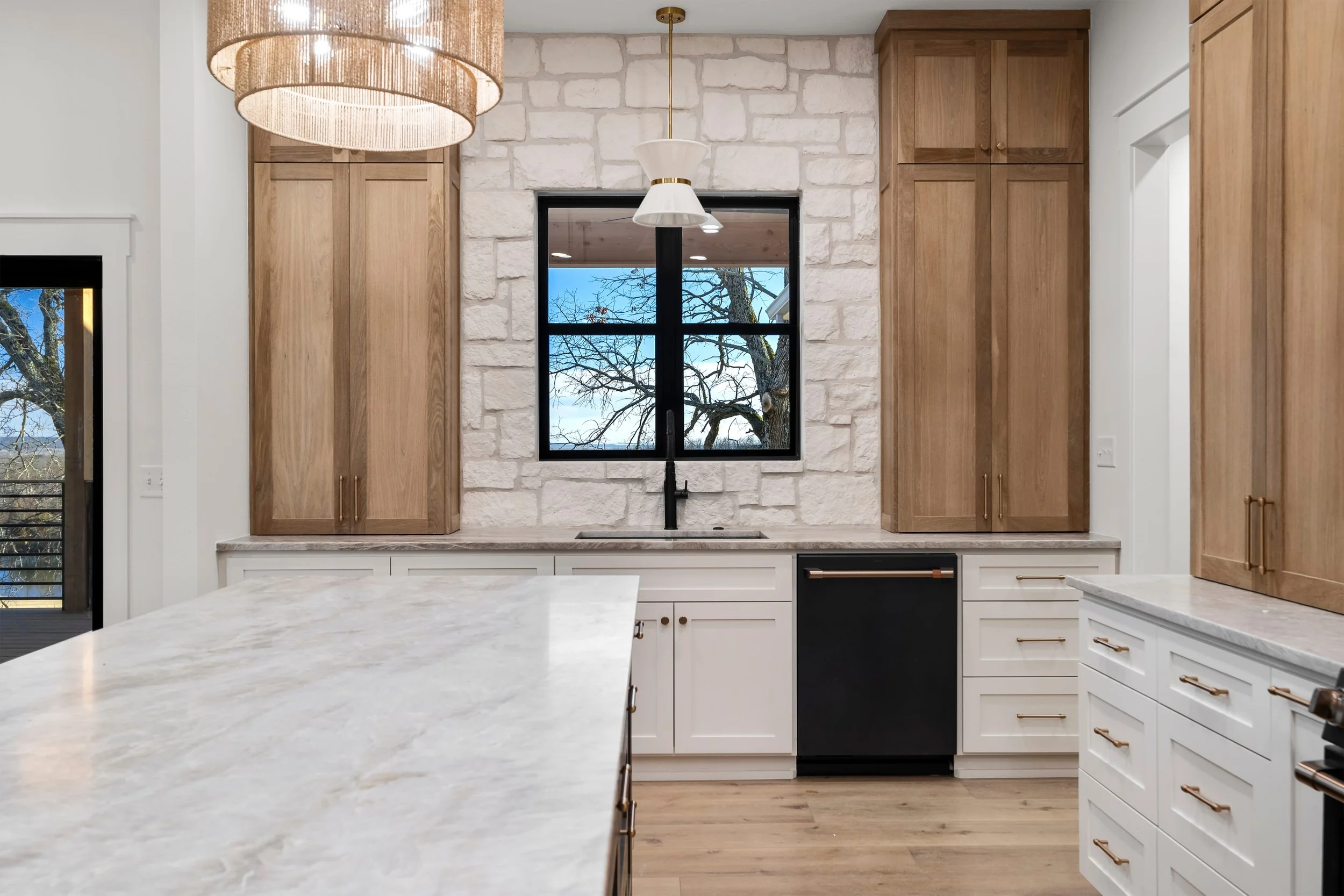 Modern kitchen with white and wood cabinetry, a black window and faucet, a marble island, and a stone wall backdrop, with a view of trees outside.