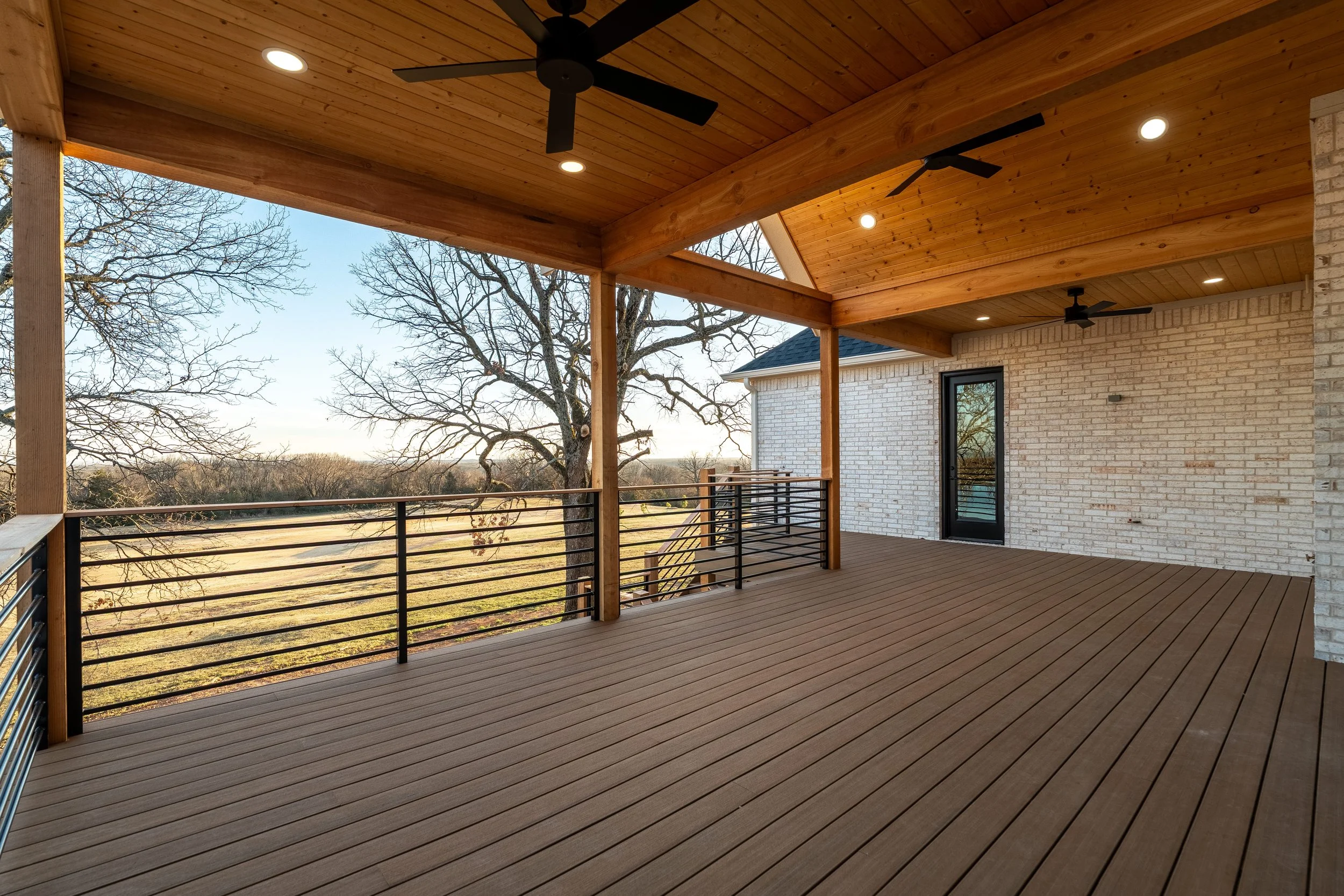 Covered wooden balcony overlooking a natural landscape with leafless trees and open fields, attached to a house with brick exterior and black door.