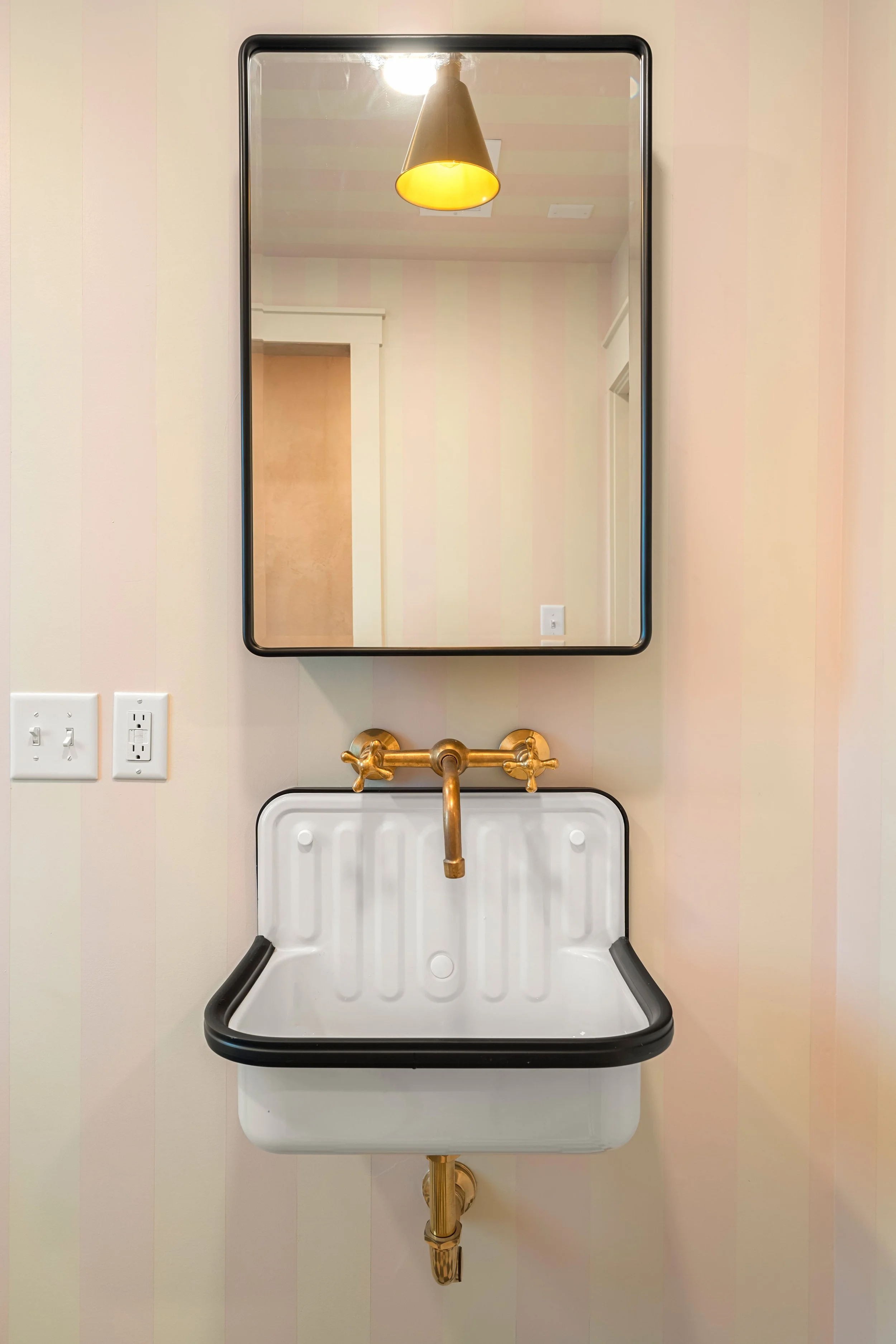 A wall-mounted, vintage-style white sink with a black rim, brass fixtures, and a matching brass pipe underneath. Above the sink is a rectangular mirror with a black frame, reflecting a ceiling light with a yellow shade and a part of a doorway, with a