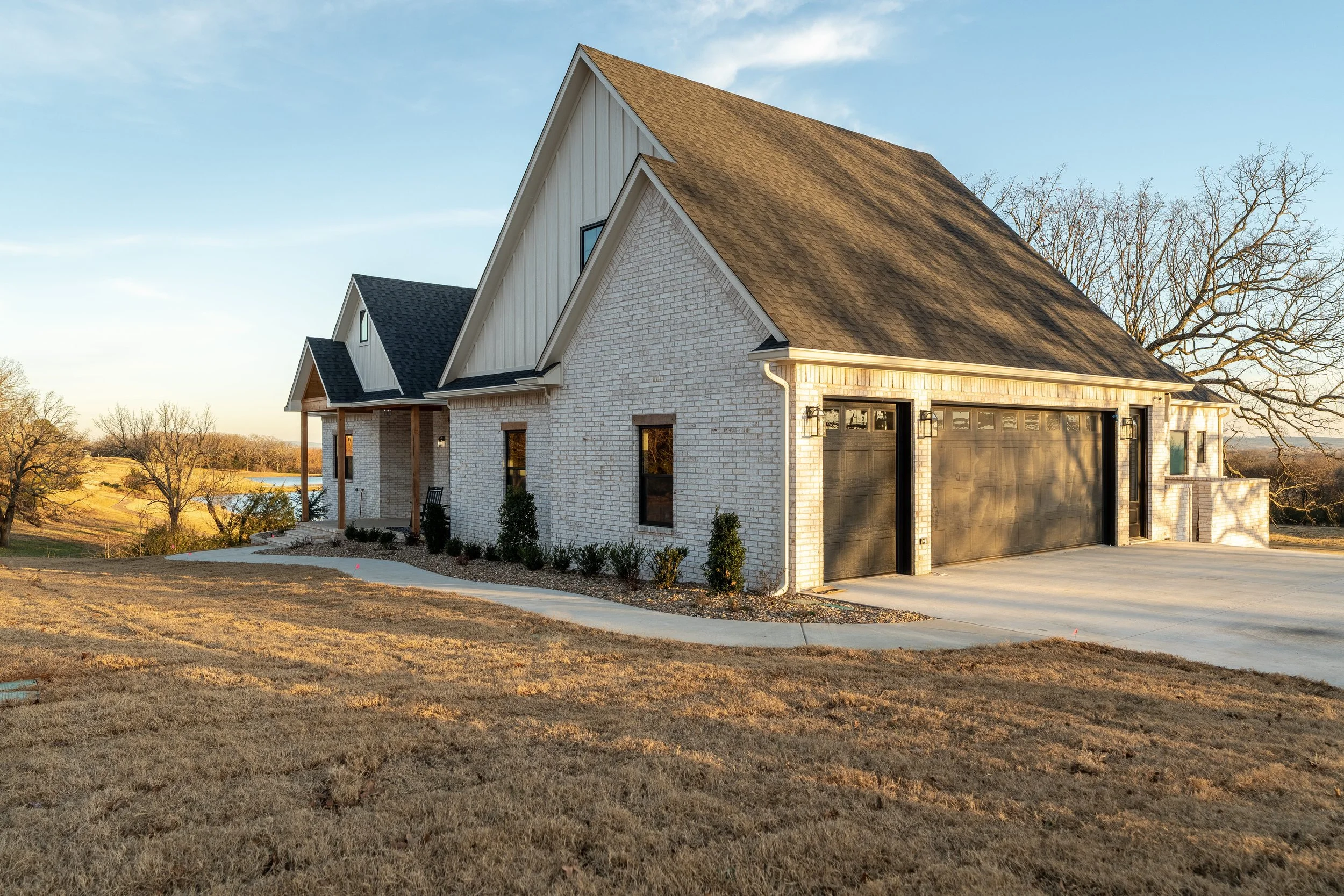 A modern two-story house with white brick exterior, black garage doors, and a pitched roof, set against a rural landscape with a pond and trees in the background.
