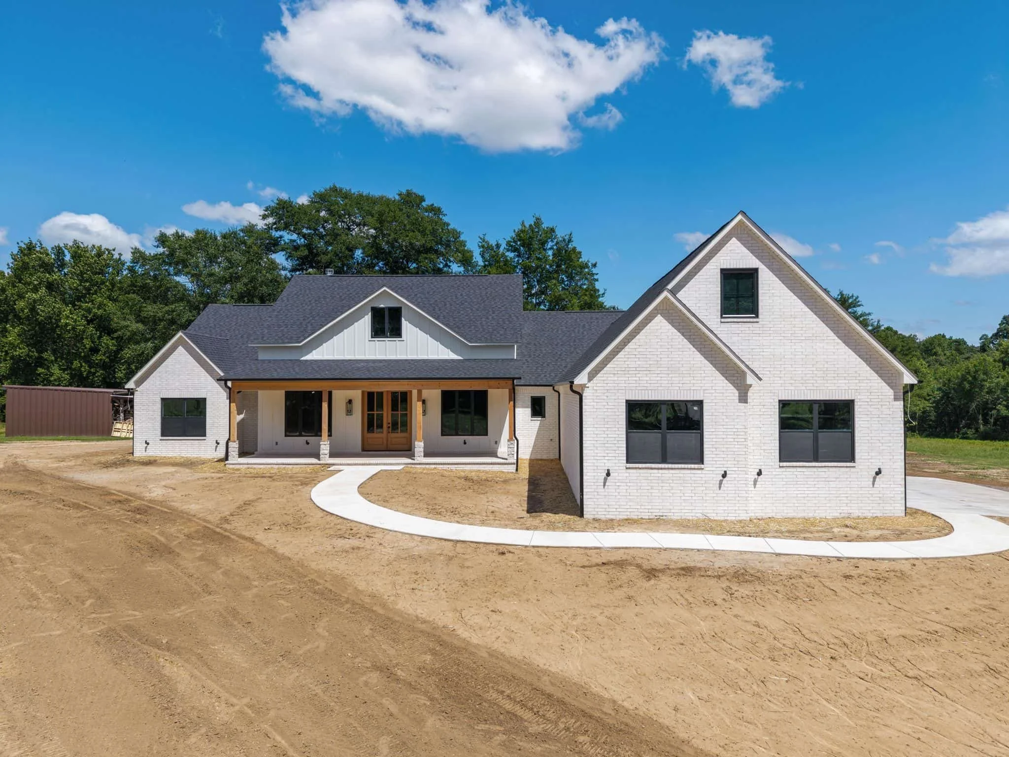 Front view of a white brick house under construction with a curved concrete walkway leading to the front door, surrounded by dirt and construction site area, with trees and a partly cloudy blue sky in the background.
