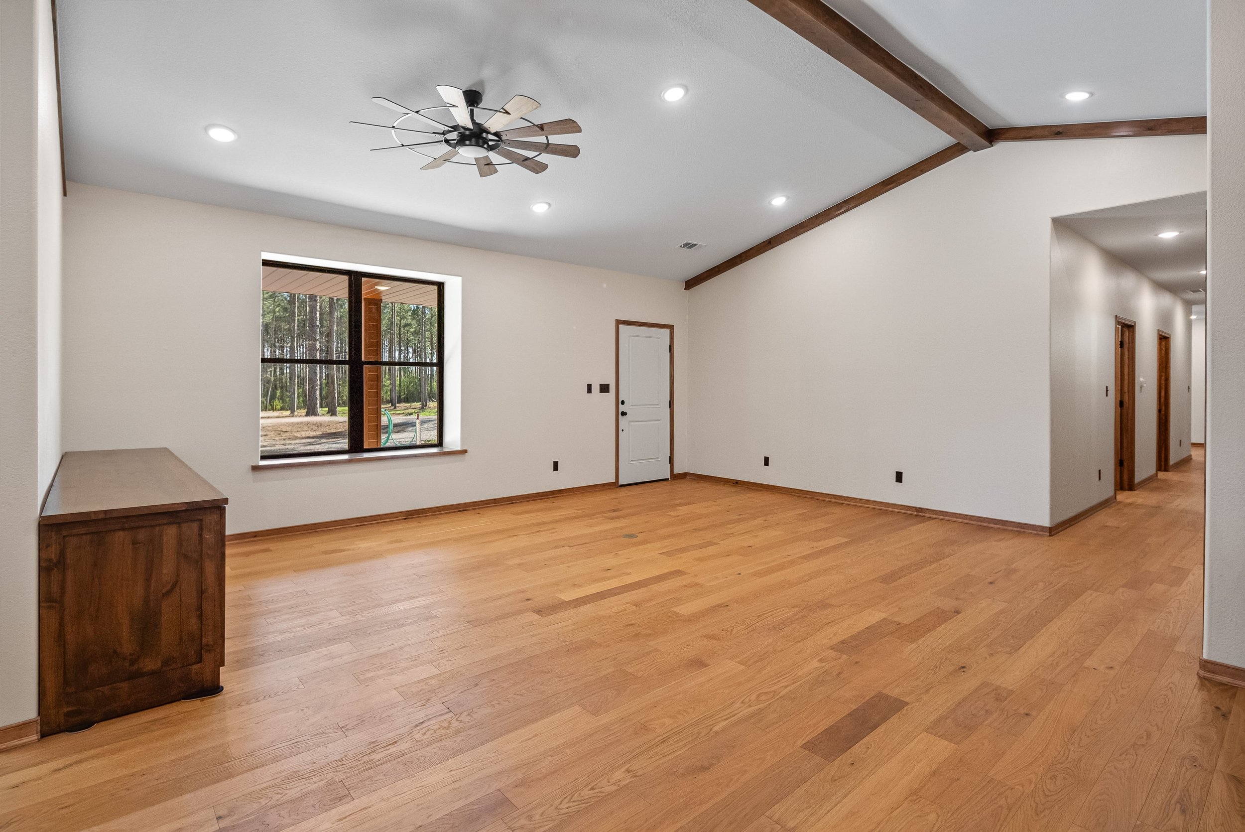 Empty living room with hardwood floors, white walls, a window showing a wooded outdoor area, a wooden ceiling fan, and recessed ceiling lights.