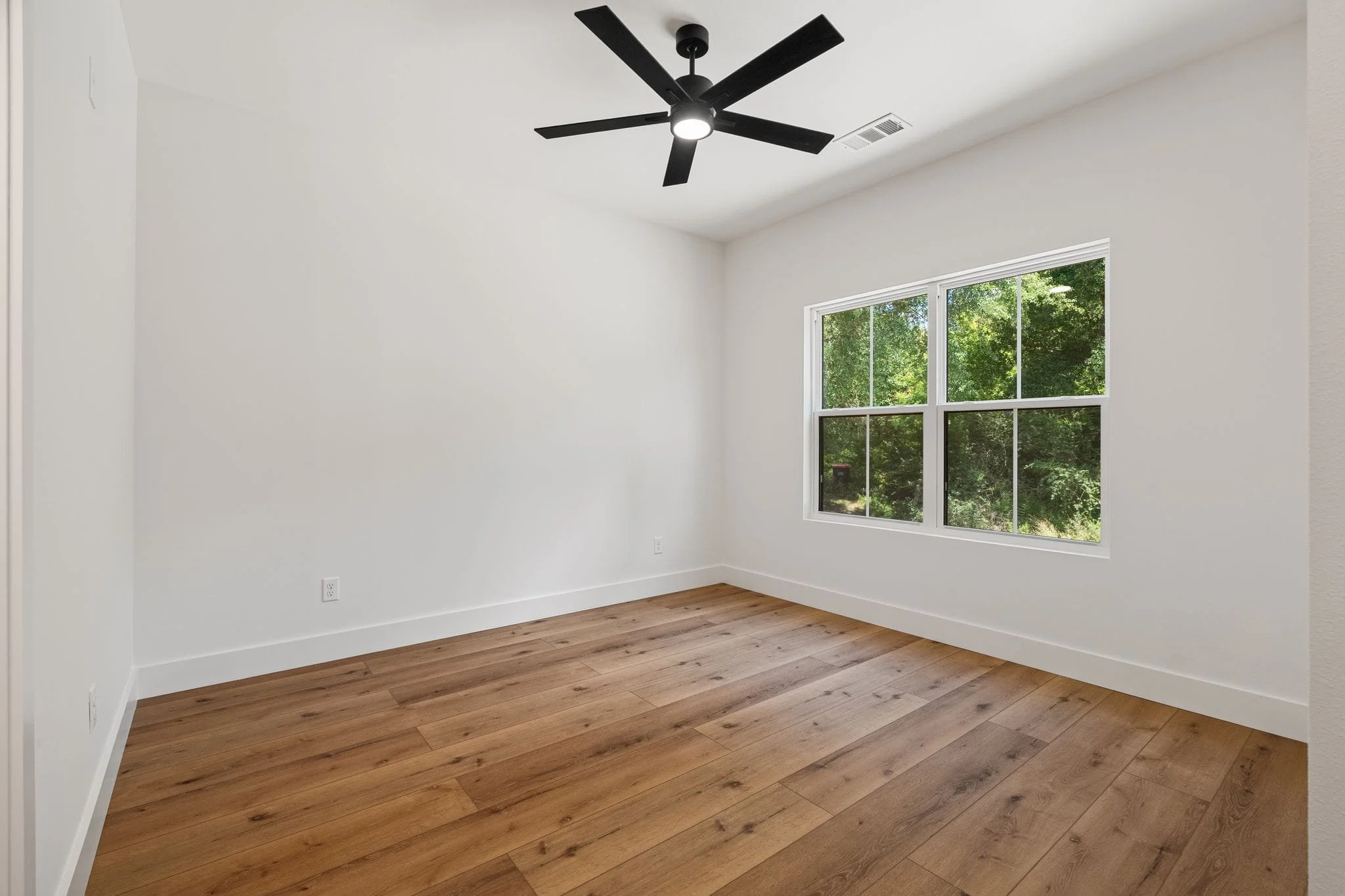 Empty room with white walls, hardwood flooring, a large window showing green trees outside, and a black ceiling fan.