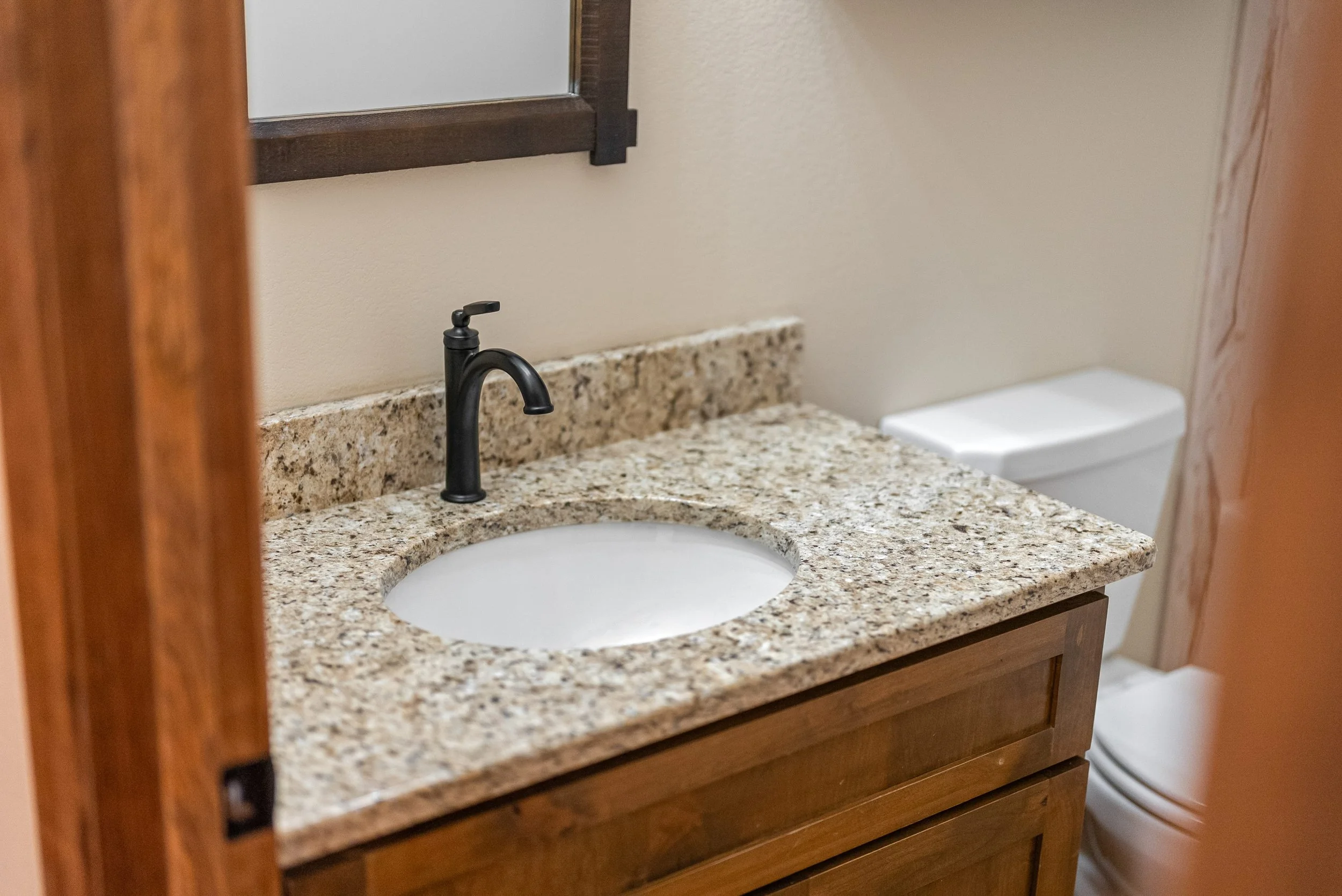 Bathroom vanity with a granite countertop, black faucet, and undermount sink. A mirror is partially visible above the countertop, and a white toilet is in the background.