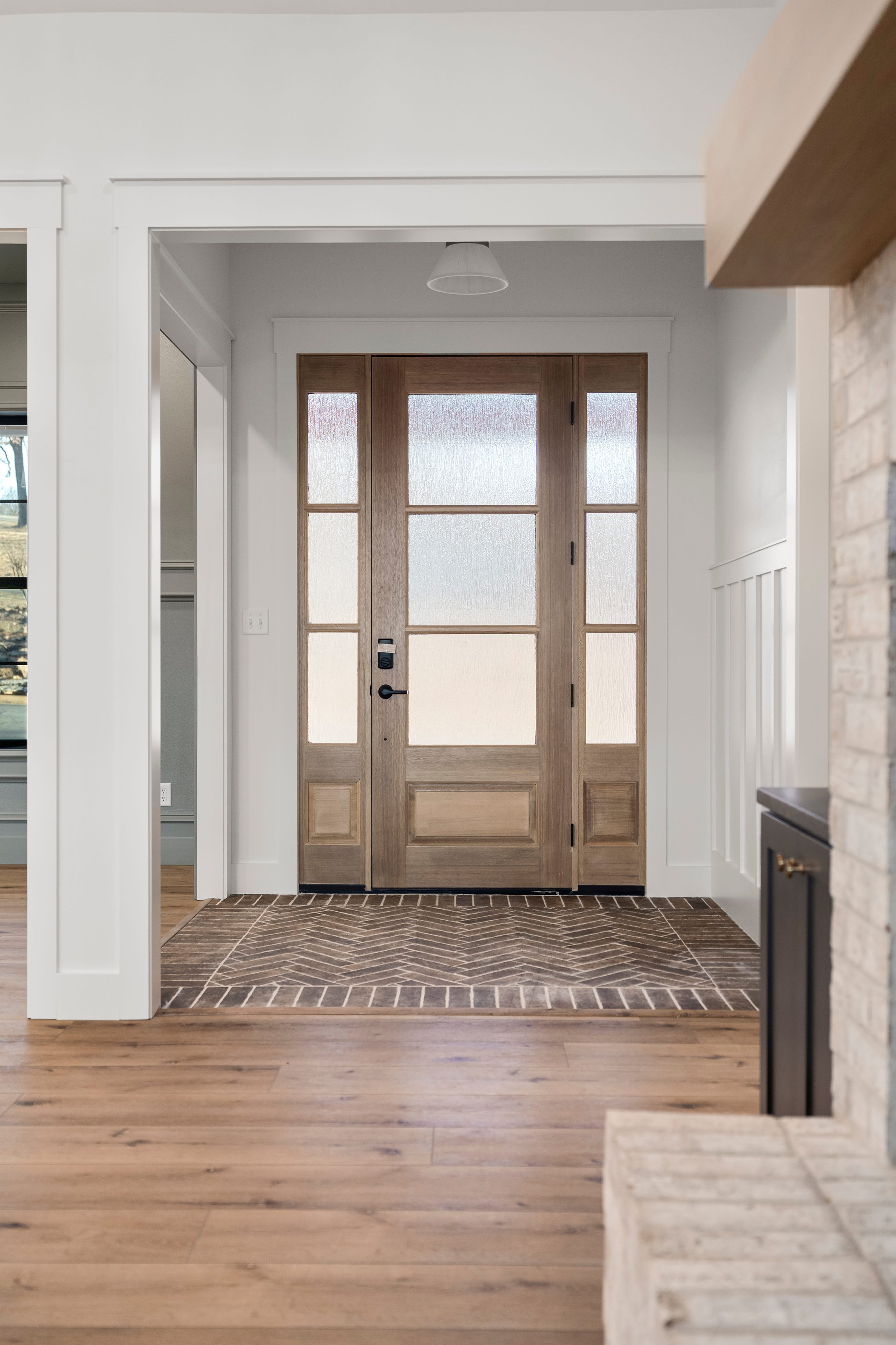 Wooden front door with glass panels in a house entryway, with a brick and wood floor, white walls, and some ceiling lighting visible.