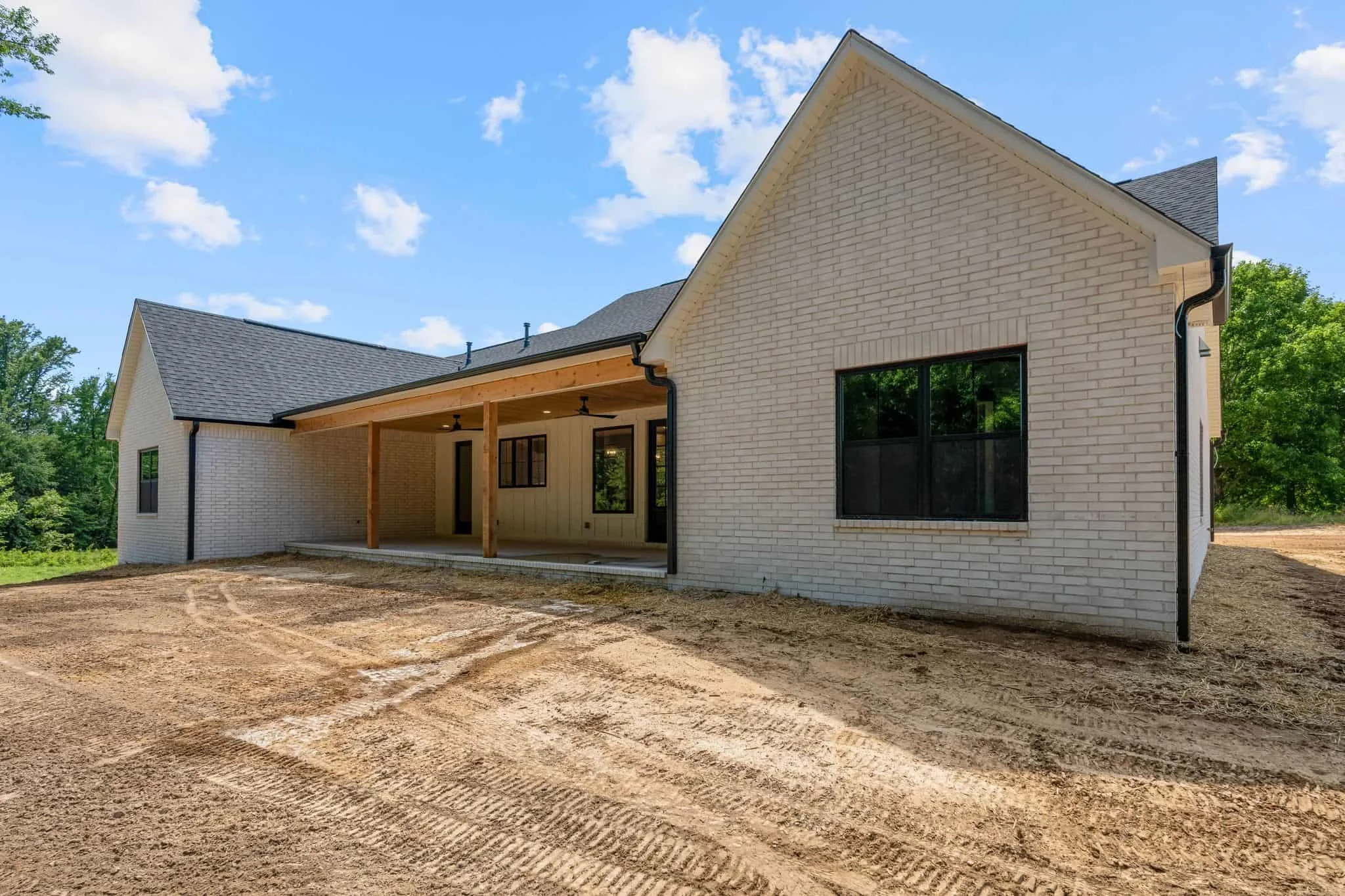 Newly constructed house with white brick exterior, large black window, and covered back porch with wooden beams, set against blue sky with clouds and surrounded by trees, with bare dirt in the foreground.