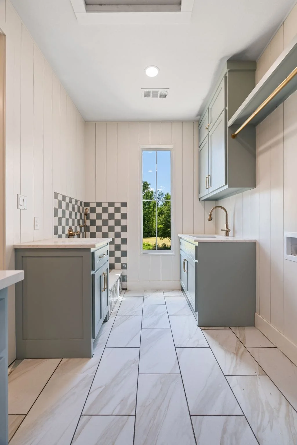 Clean laundry room with light gray cabinets, a window showing green trees outside, white walls with vertical paneling, a tiled floor, and a checkered backsplash area behind a utility sink.