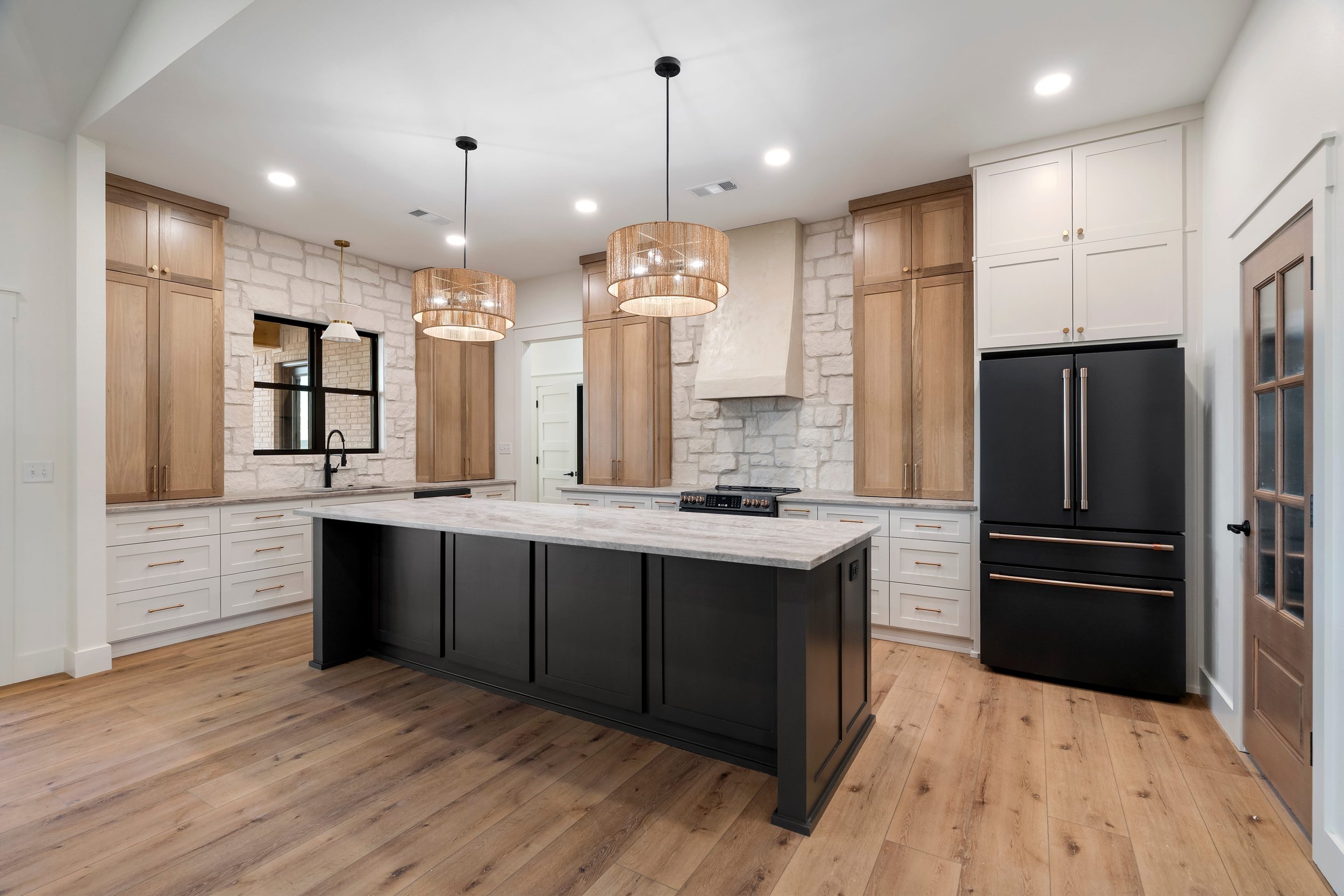 Modern kitchen with black island, white cabinets, wooden accents, stone wall, pendant lights, black refrigerator, wooden door, and hardwood floors.