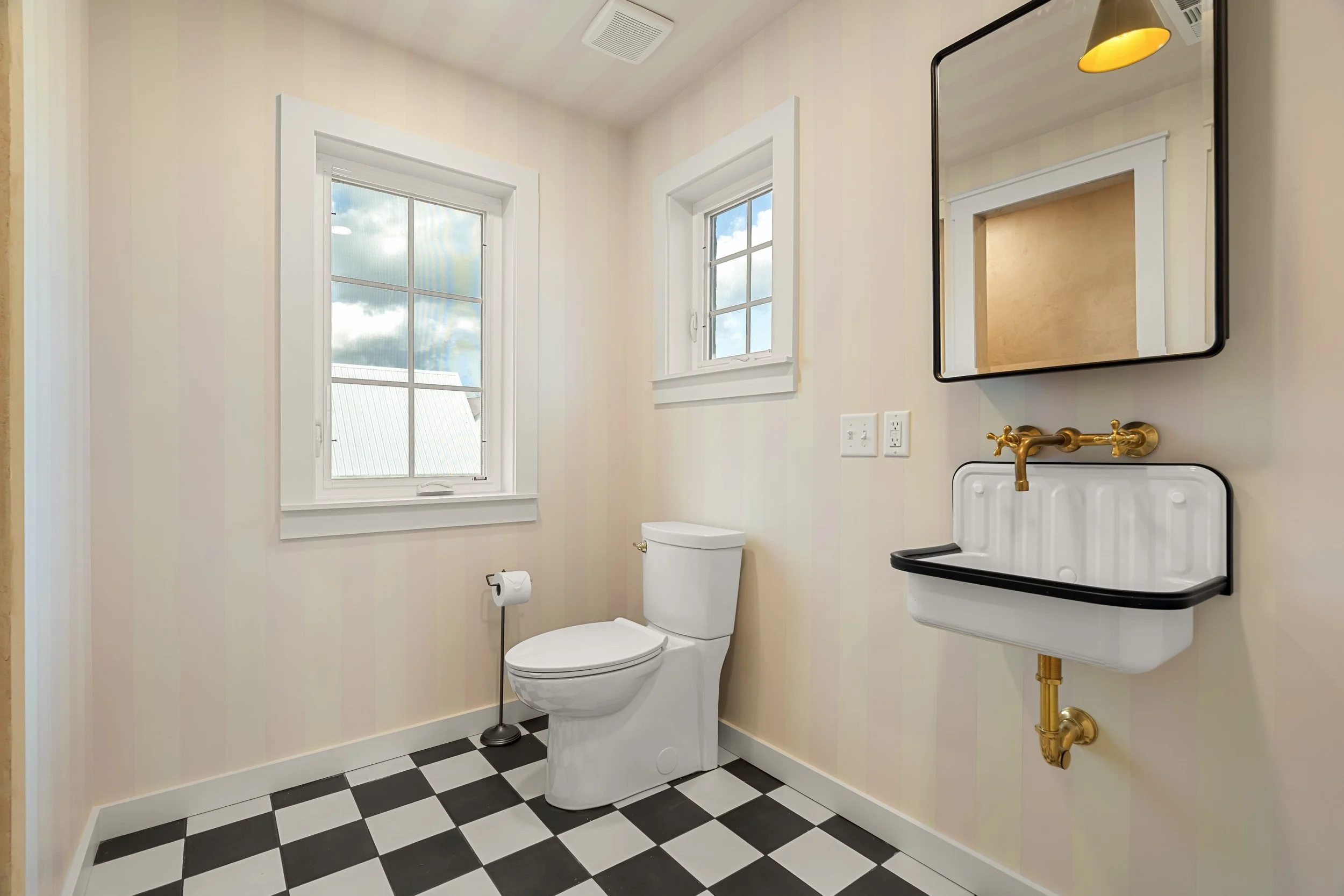 Small bathroom with a white toilet, a small white sink with black trim, a mirror with black frame, a window showing a cloudy sky, and black-and-white checkered floor.