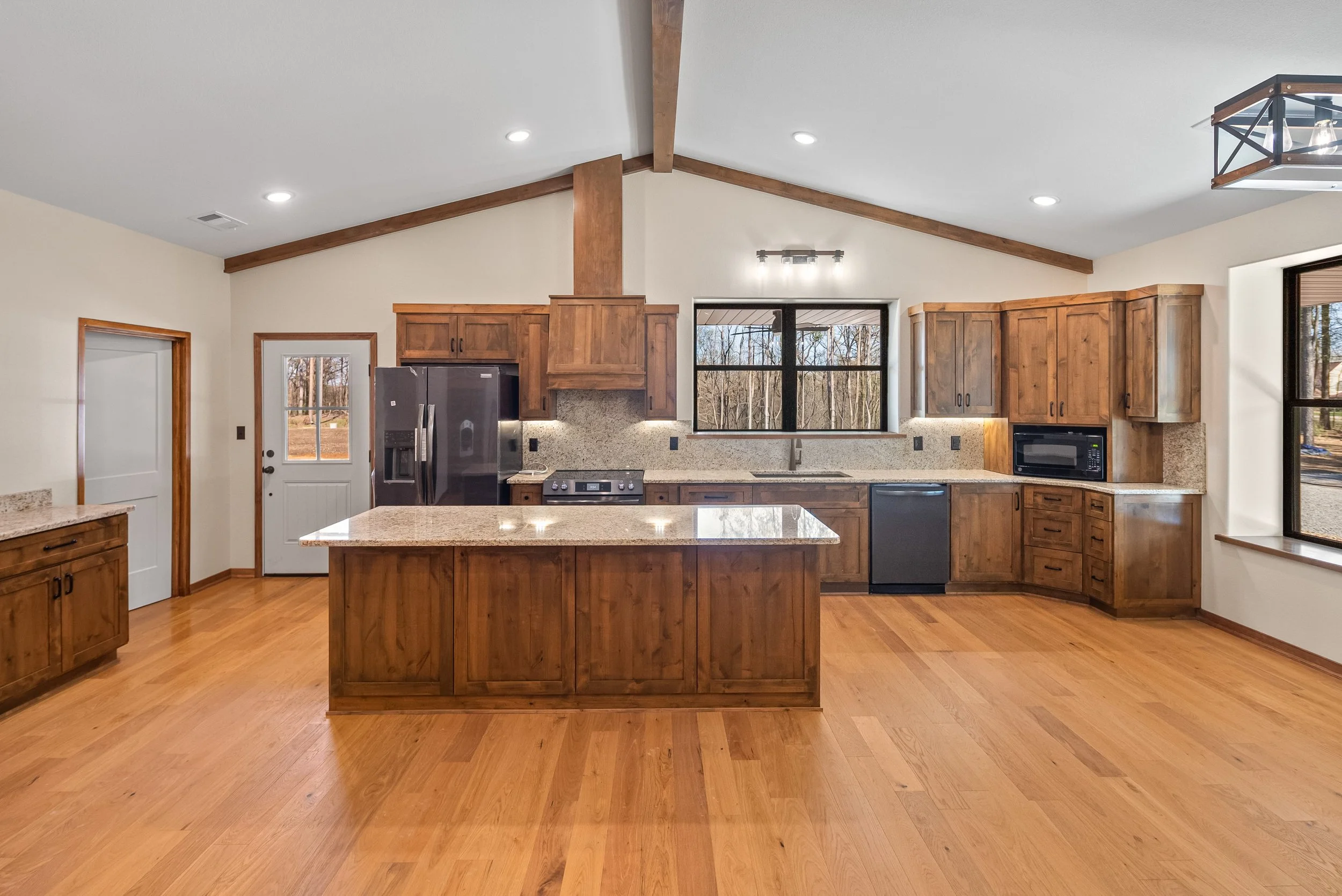 Spacious kitchen with wooden cabinets, granite countertops, island, stainless steel appliances, and hardwood floors with large window showing outdoor trees.