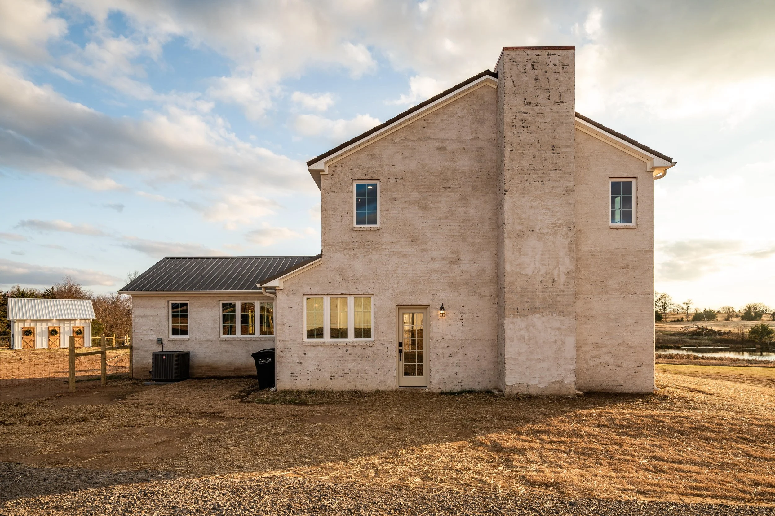 A two-story house with light-colored brick exterior, multiple windows, and a door with glass panels, set on a dirt yard under a partly cloudy sky.