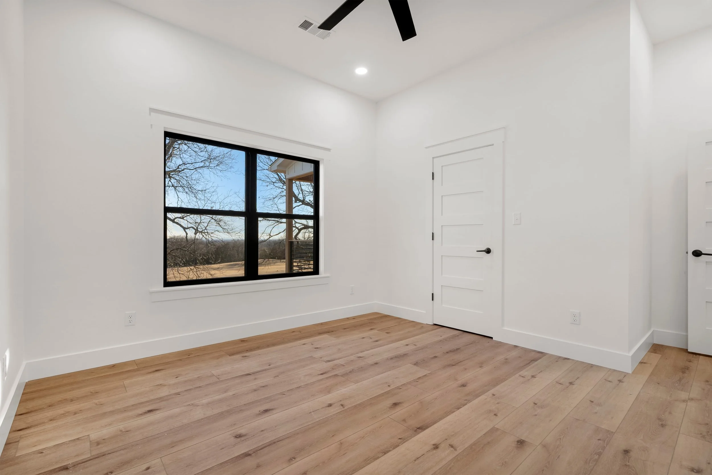 Empty bedroom with light wood flooring, white walls, a large black-trimmed window showing a scenic outdoor view, and two modern white panel doors with black handles.