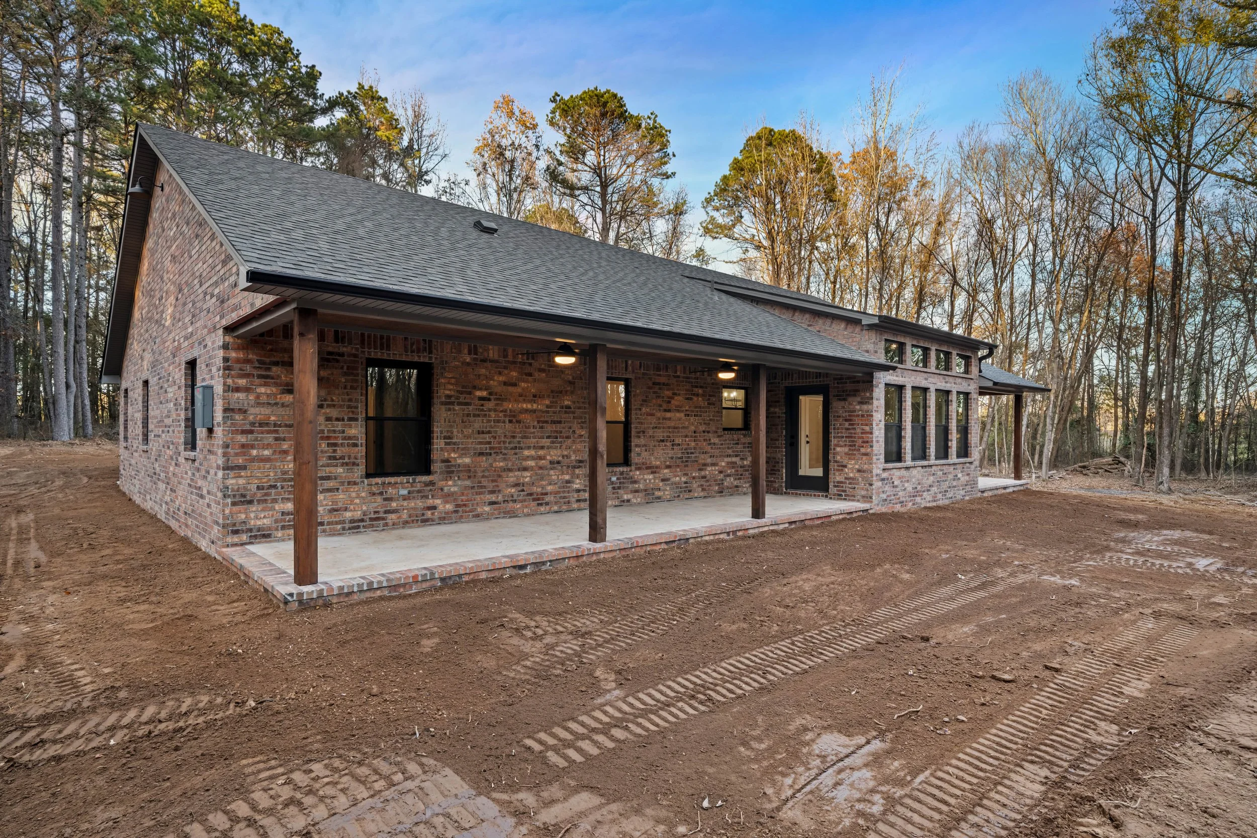 Newly built brick house with a covered patio, black door, and large windows, situated in a wooded area with dirt surroundings.
