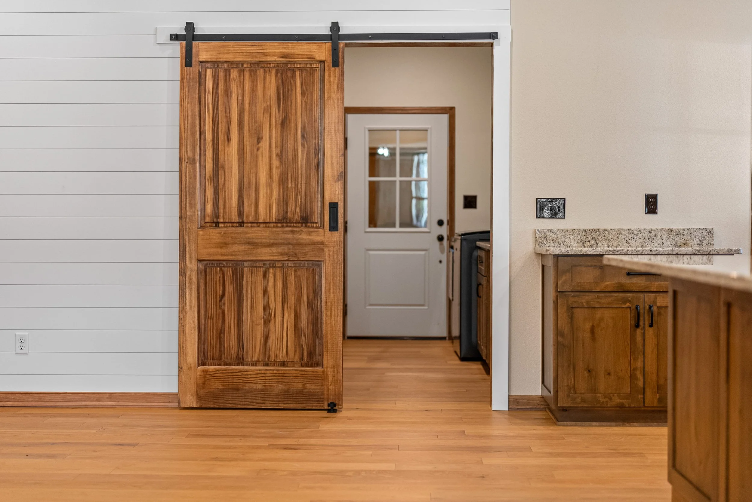 Interior of a home featuring a sliding wooden barn door on a black metal track, leading to a kitchen area with wooden cabinets and a white door with a window in the background.