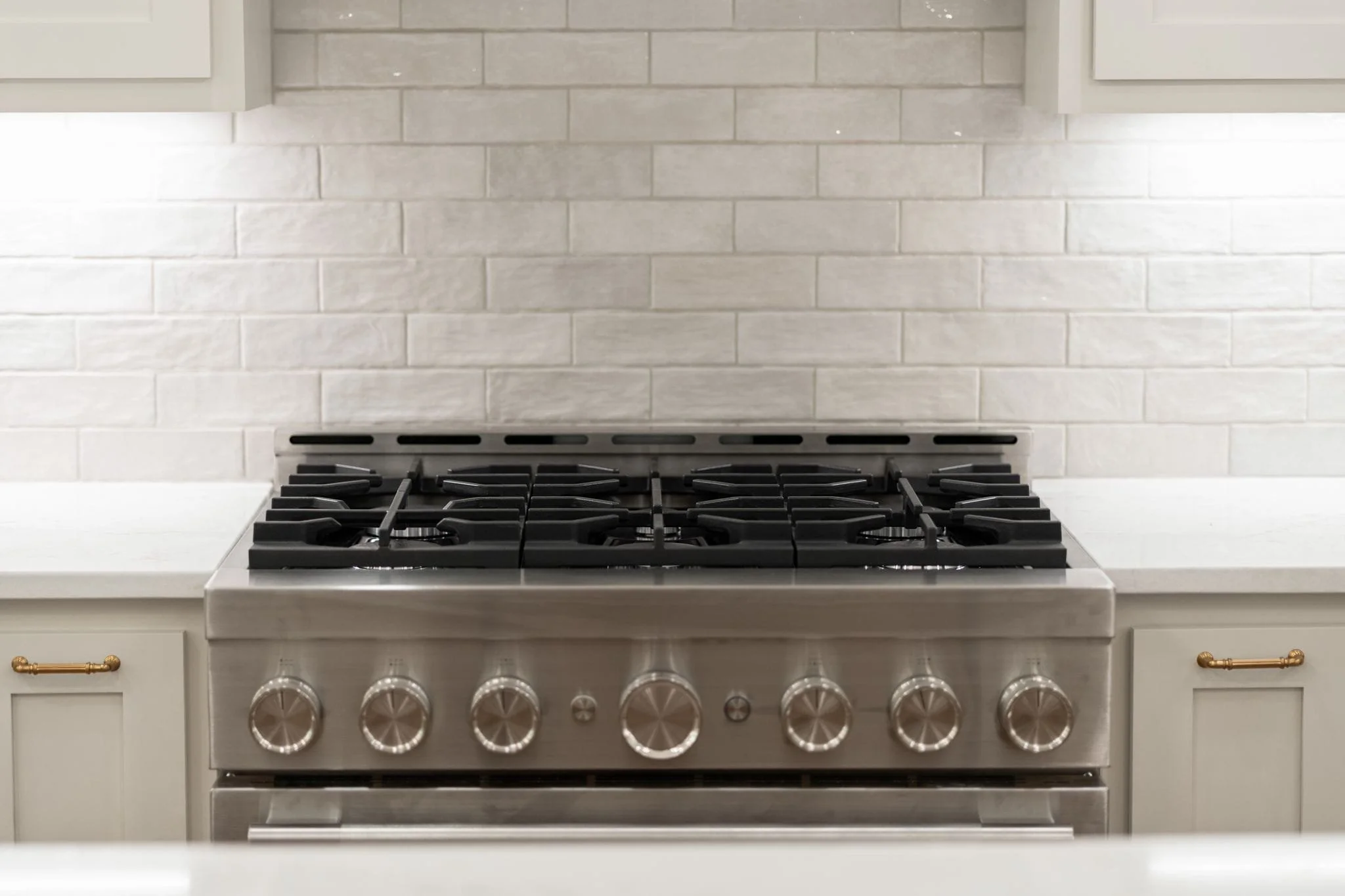Stainless steel gas stove with black burners and control knobs in a kitchen with white cabinetry and a light gray tile backsplash.