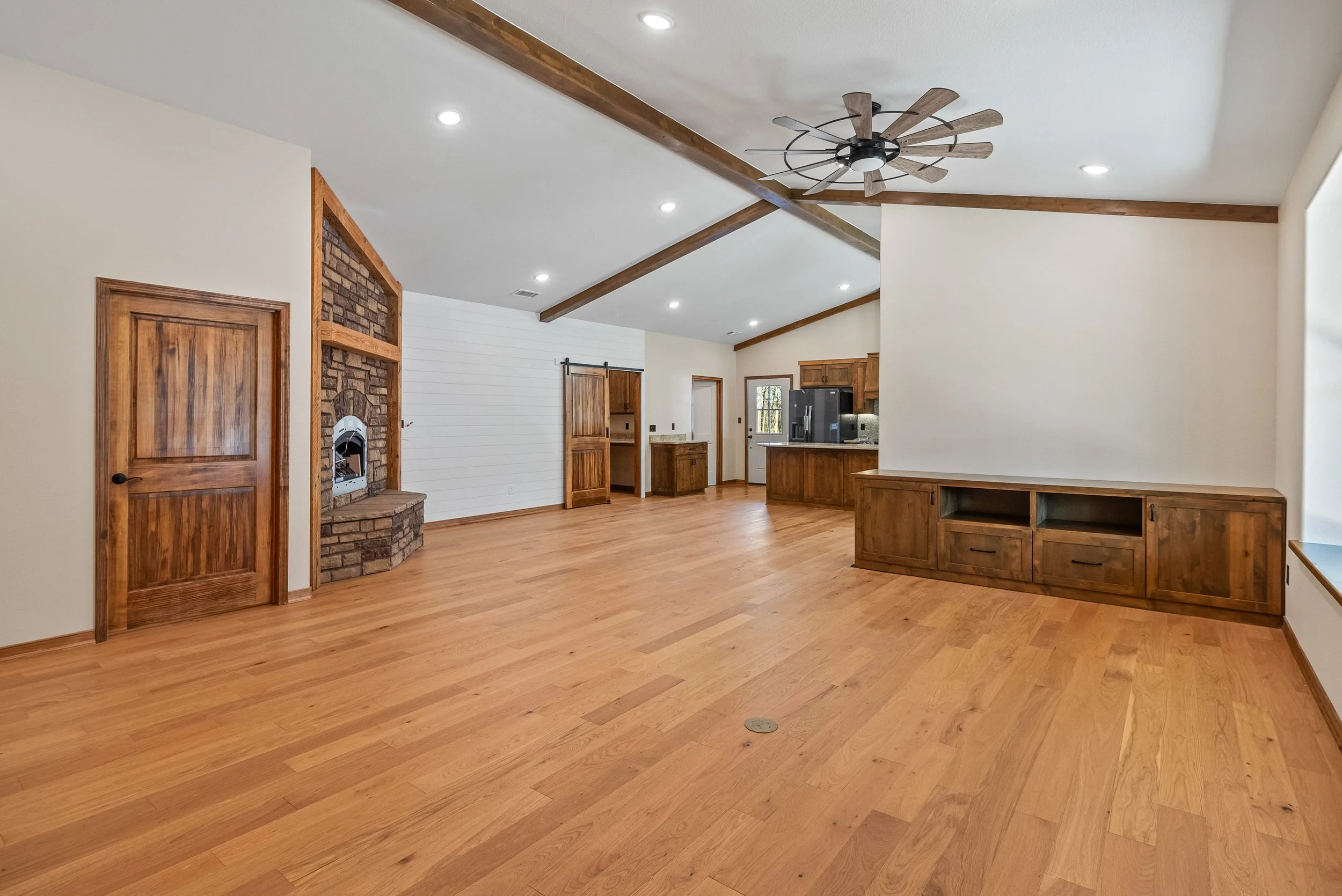 Empty living room with hardwood floors, cream walls, wood trim, a ceiling fan, and a brick fireplace. The space opens to a kitchen area with wooden cabinets and black appliances.