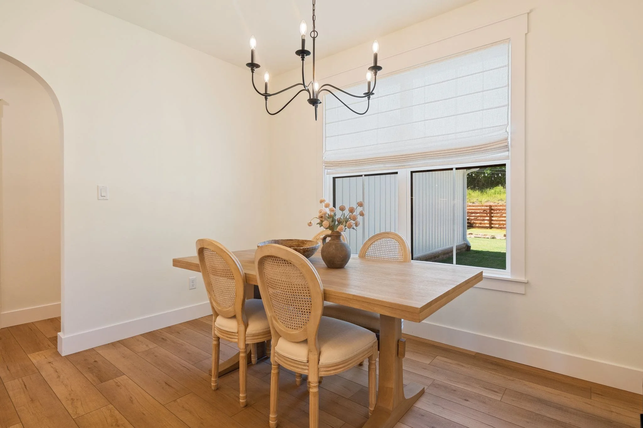 Dining nook with wood table, cane-back chairs, black chandelier, and large window overlooking the yard.