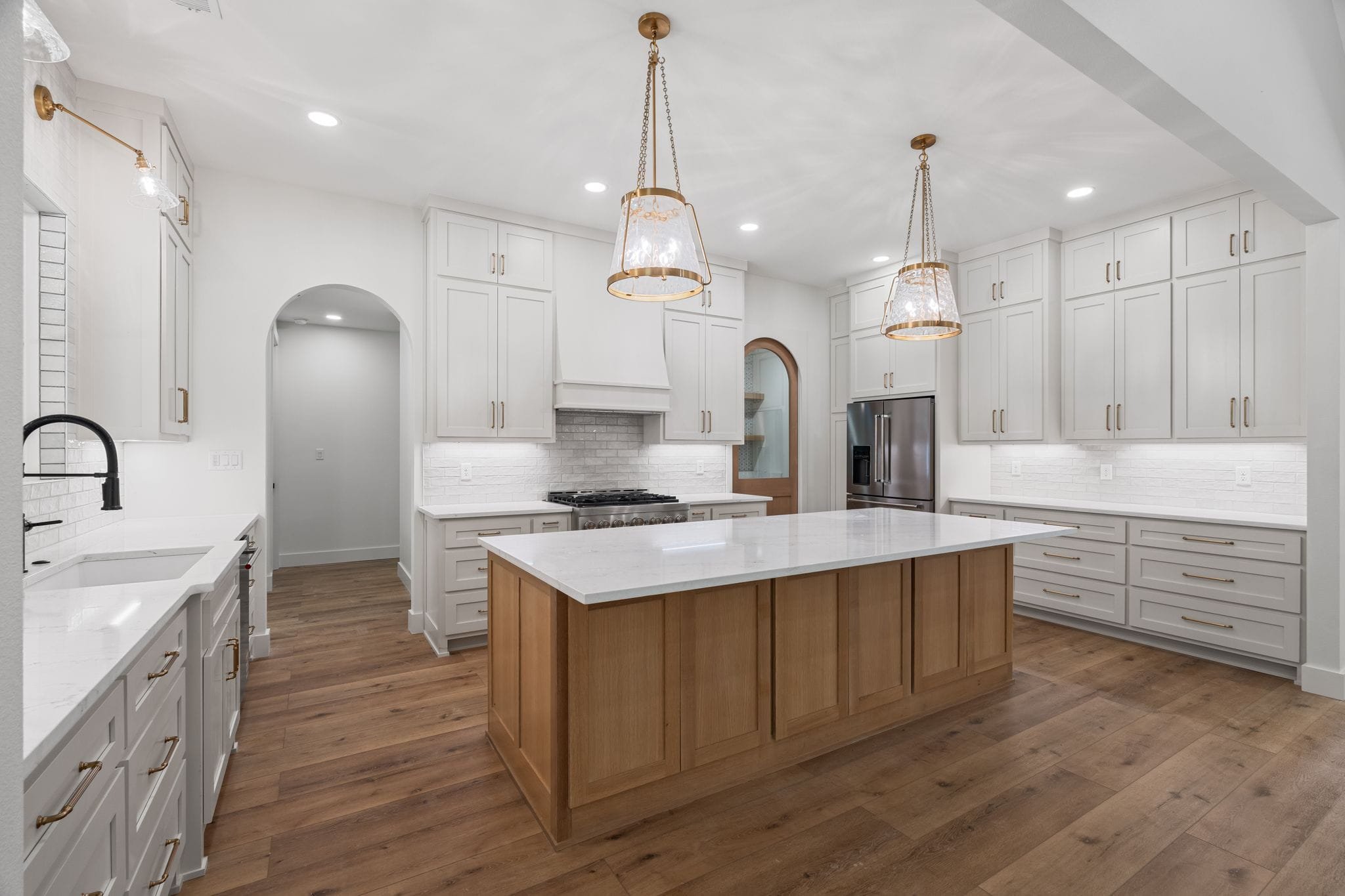 Modern white kitchen with wooden island, gold hardware, pendant lights, and hardwood flooring.