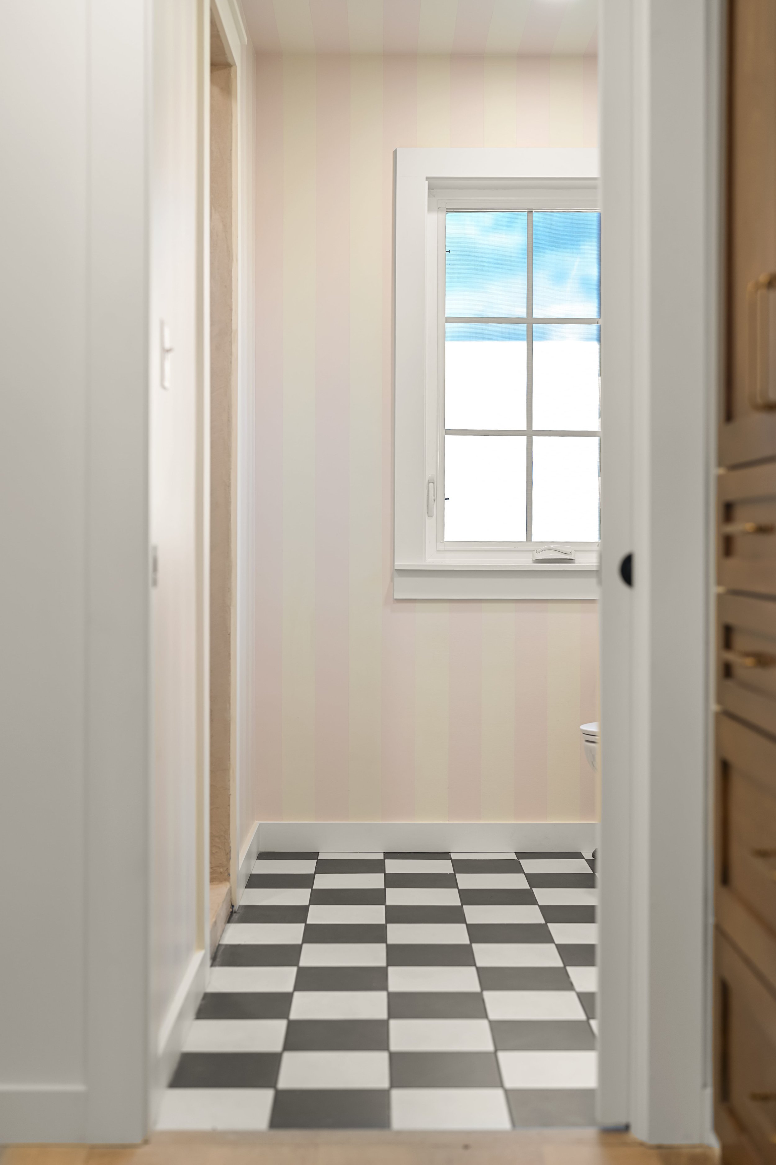 View of a checkered black-and-white tiled floor, with a window showing blue sky and clouds, and part of a bathroom visible through an open door.