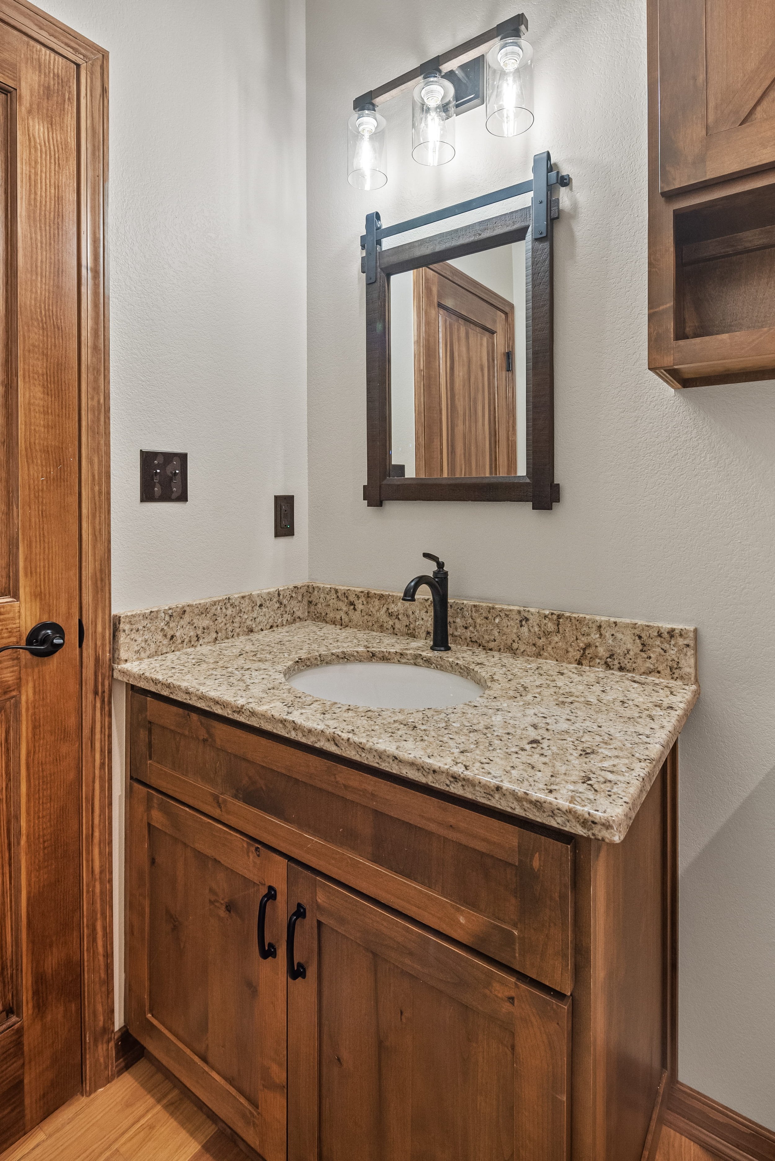 Bathroom vanity with granite countertop, a round undermount sink, black faucet, rectangular mirror with dark wood frame, wooden cabinet, and wall-mounted light fixture with three clear glass shades.