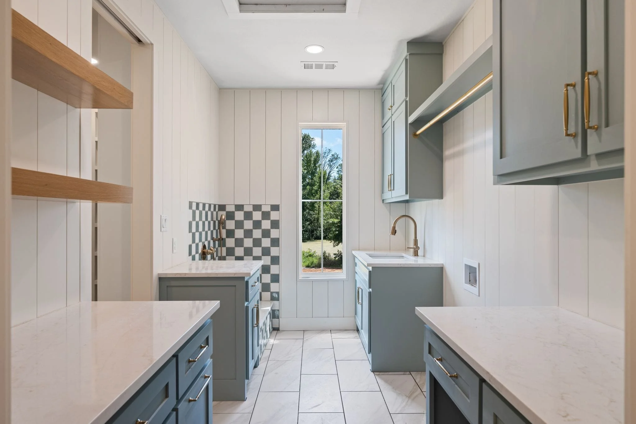 Interior view of a modern kitchen with white walls, blue cabinets with brass handles, marble countertops, and a window showing greenery outside. There is a small section of checkered backsplash next to a window.