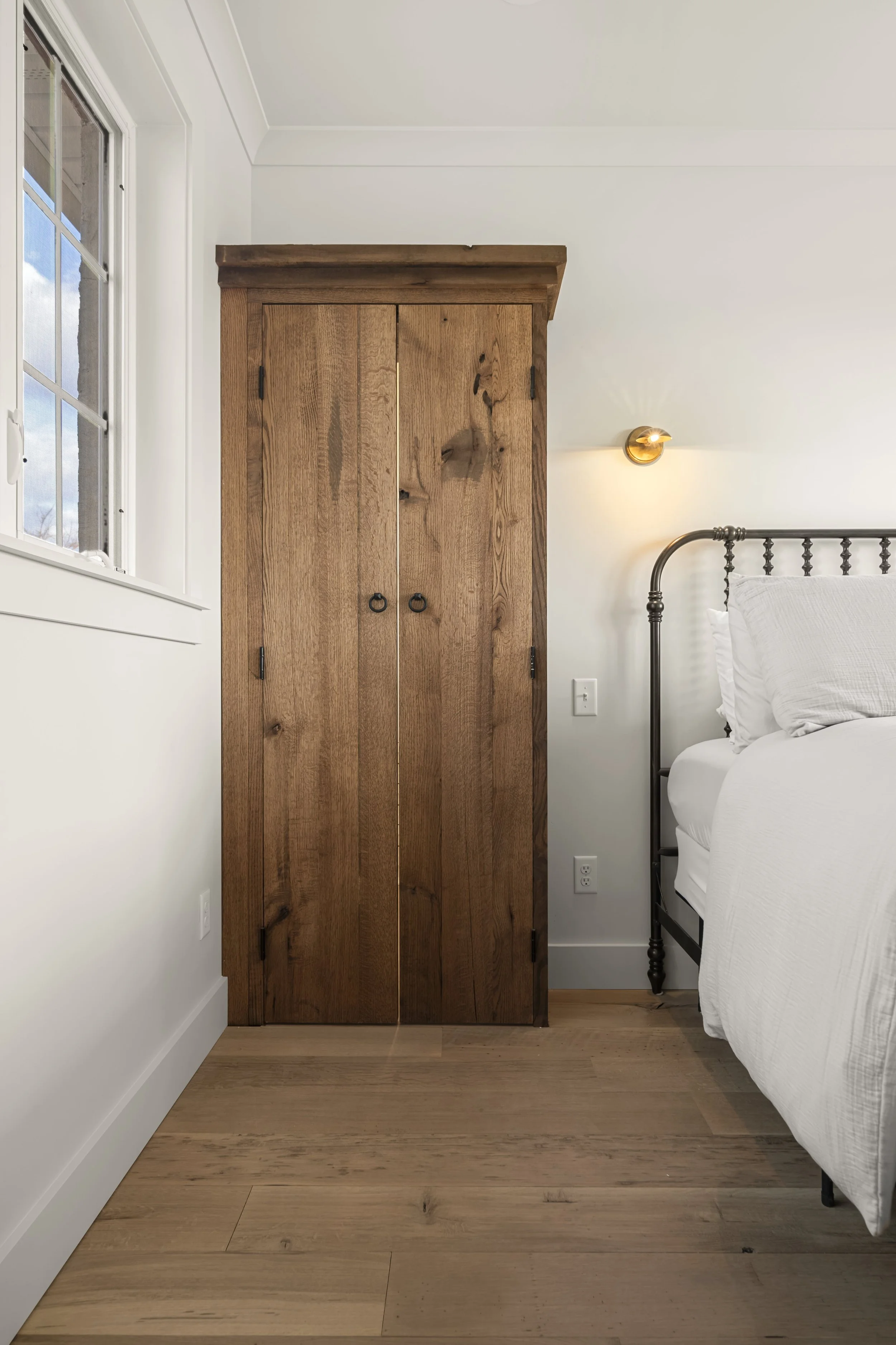 A wooden wardrobe next to a white bed with white bedding in a bedroom with a white wall, hardwood floor, a window, and a small wall-mounted light fixture.