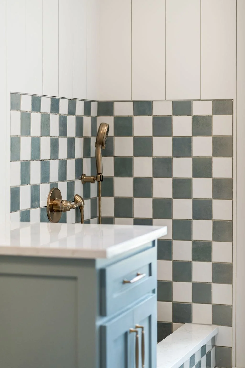 Bathroom shower with a brass handheld showerhead and matching brass fixtures, set against a wall with a checkered pattern of white and dark green tiles, adjacent to a white countertop and light blue cabinet with gold handles.