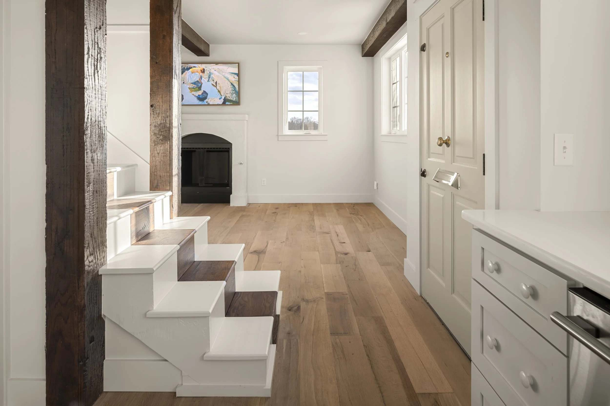 Interior view of a bright living room with wooden floors, white walls, a fireplace, three windows, and a staircase with dark wood steps.