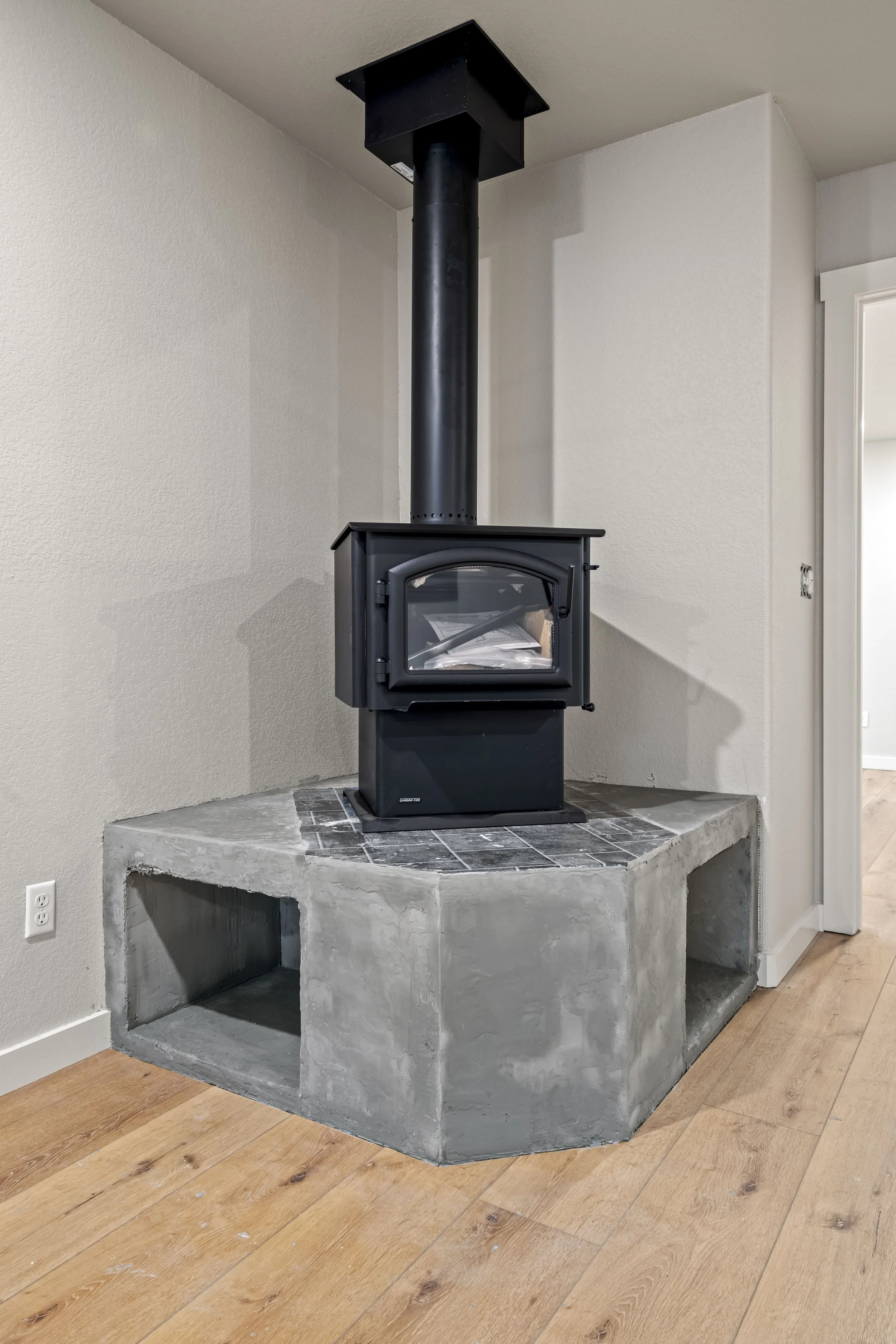 A black wood stove with a glass door is installed on a raised concrete hearth in a corner of a room, with a black chimney pipe extending to the ceiling.