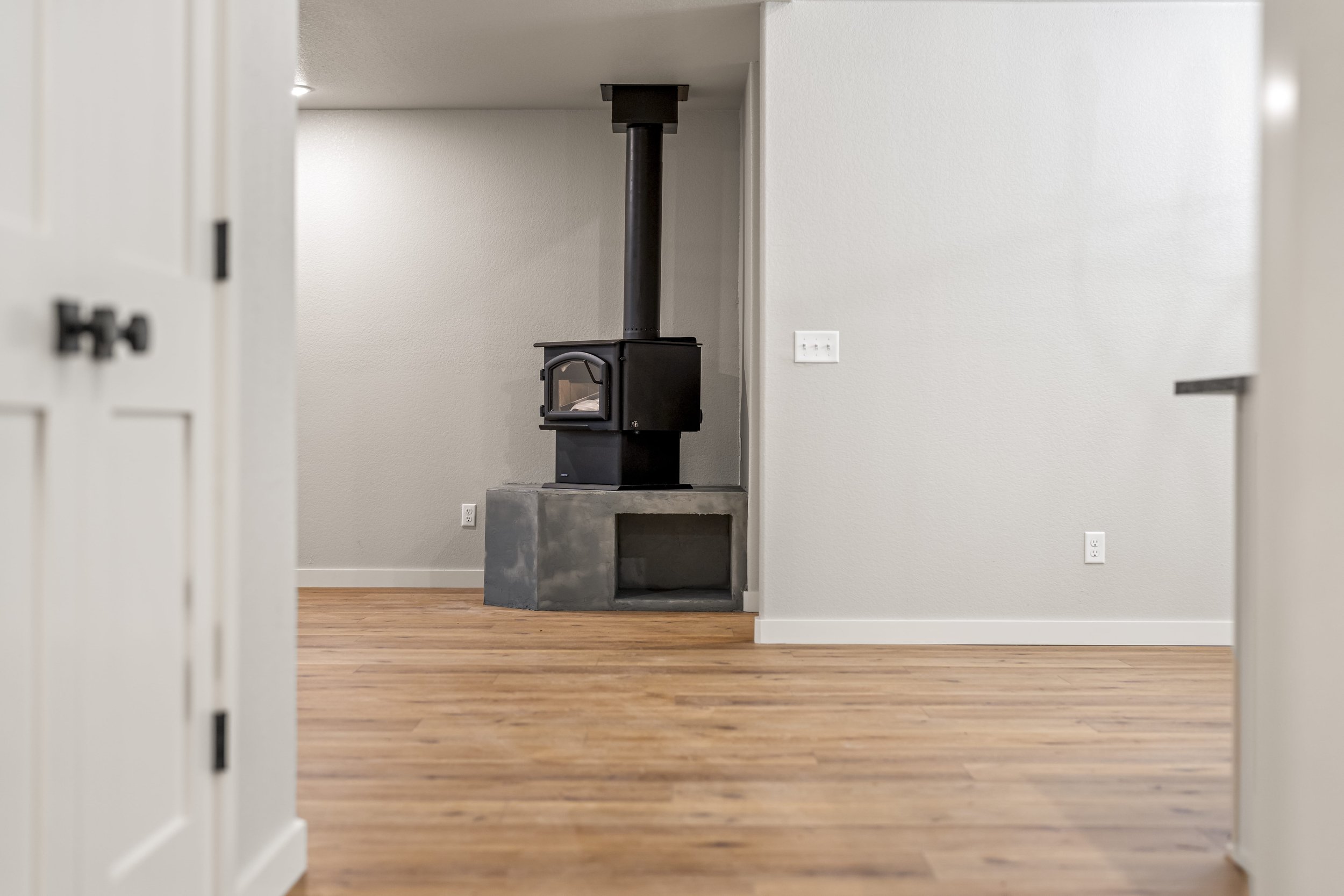 Interior view of a room with a black wood stove on a concrete platform, beige walls, hardwood floor, and a white wall outlet.