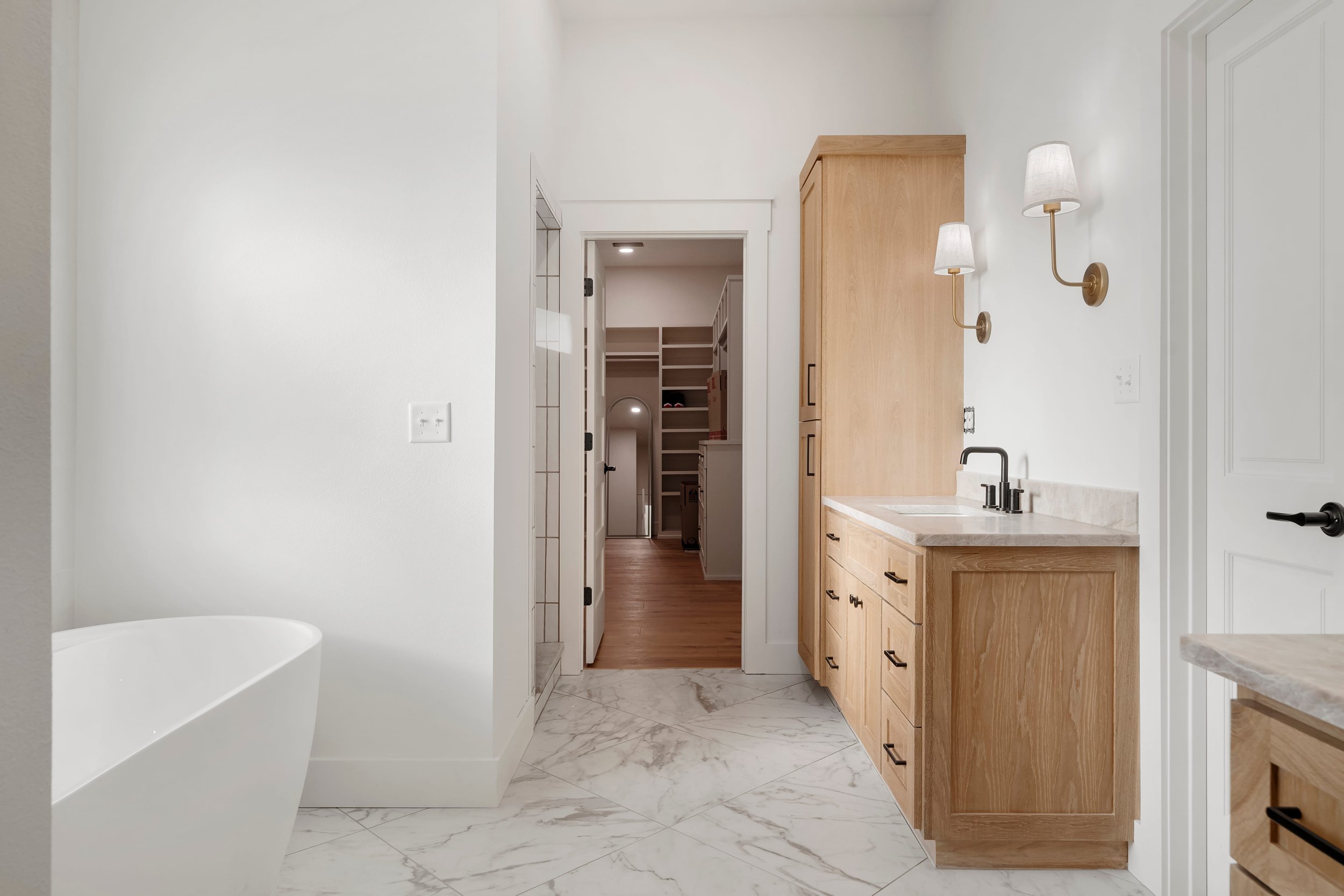 Modern bathroom with white walls, marble floors, a bathtub, wooden cabinetry, and wall-mounted lamps.