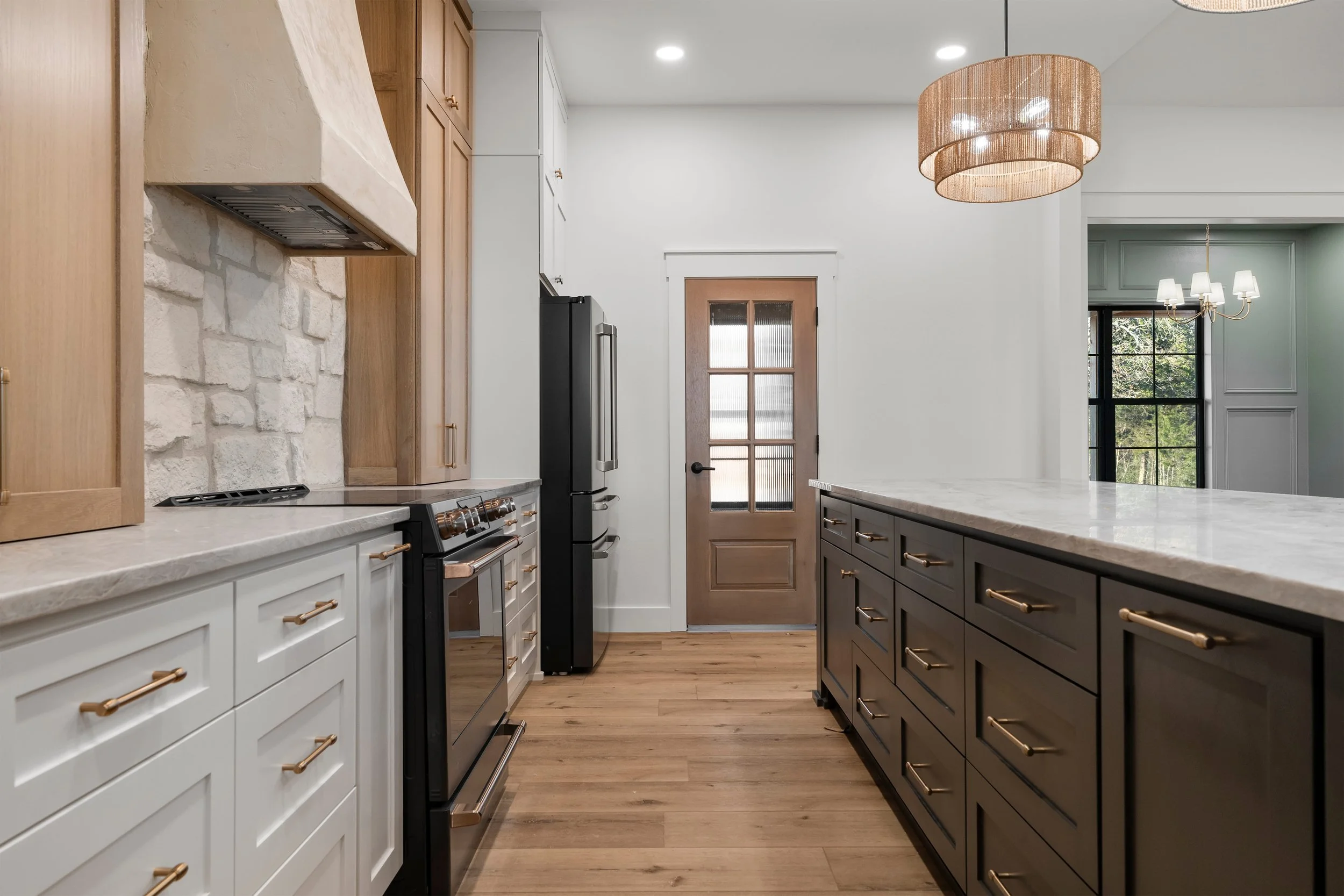 Modern kitchen with white and dark cabinetry, wood flooring, marble countertops, and pendant lighting. A wooden door with glass panels is at the end.