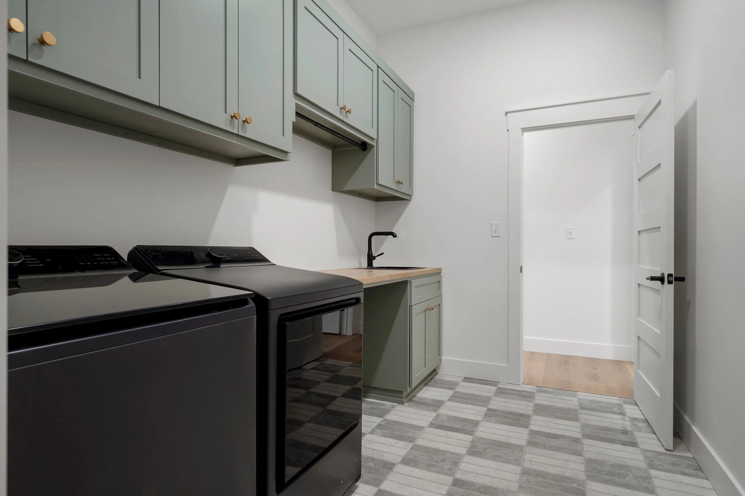 Empty laundry room with gray cabinets, black washer and dryer, sink, and patterned tile floor.