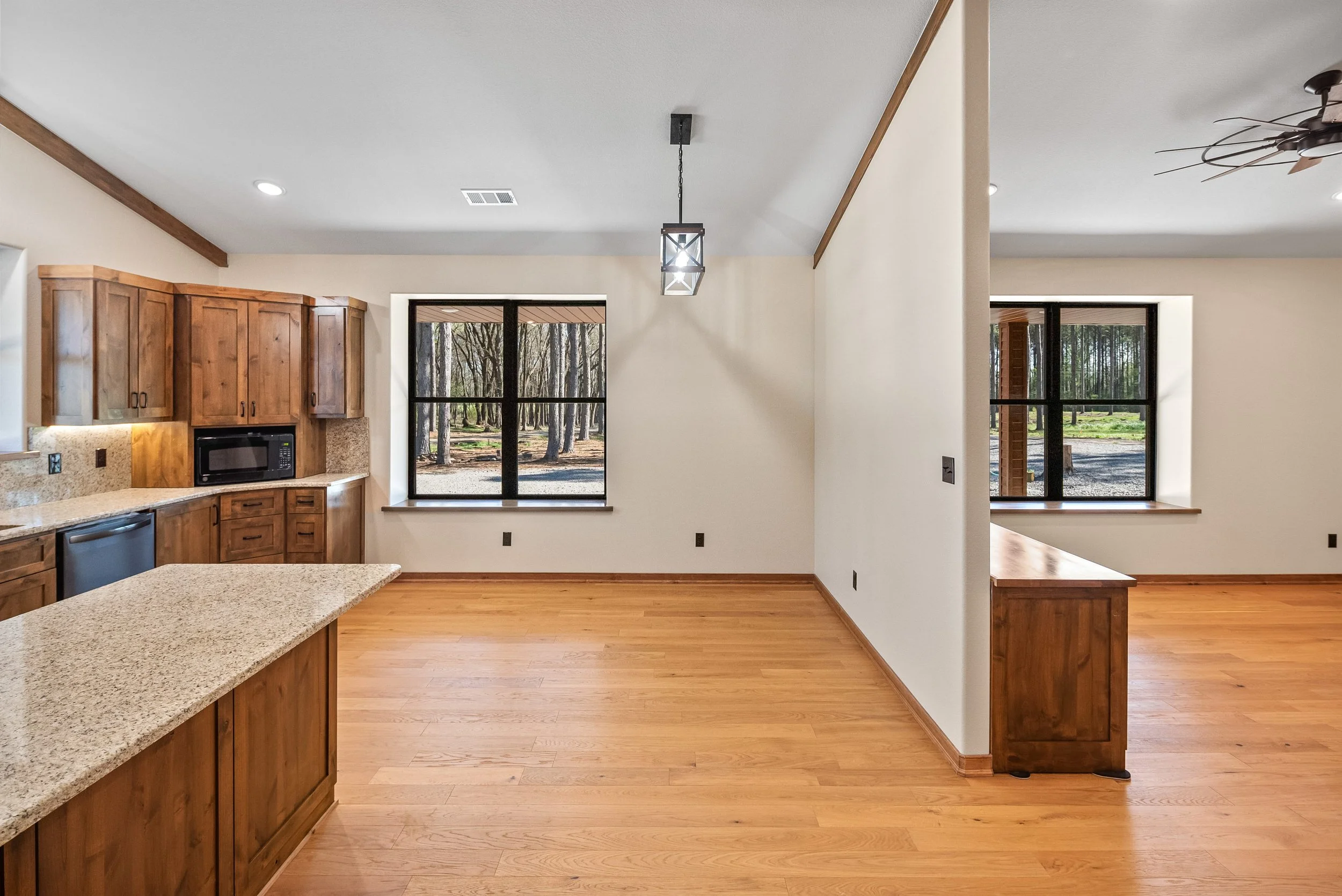 Empty kitchen and dining area with wooden cabinets, hardwood flooring, large windows showing outdoors with trees, and a ceiling fan.