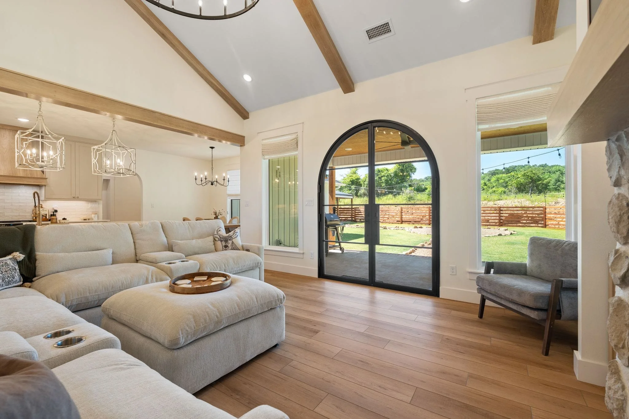 Open-concept living room with arched black patio doors, vaulted ceilings, wood beams, and backyard views.