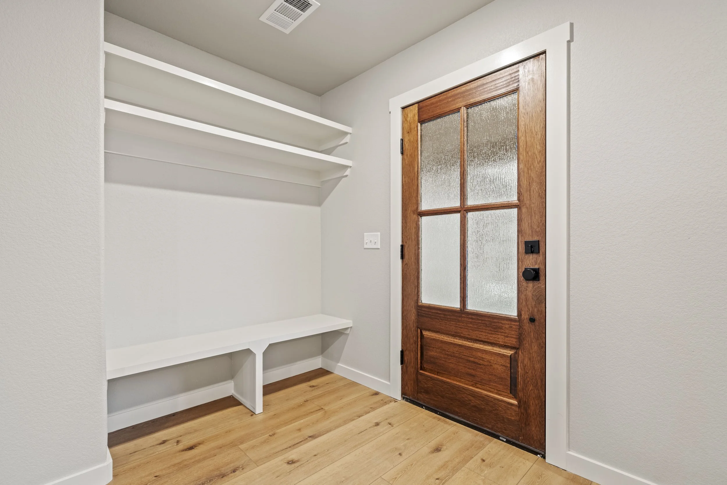 Interior of an entryway with a wooden door with glass panels on the right, white walls, built-in white shelves and bench on the left, wood floor, and ceiling vent.