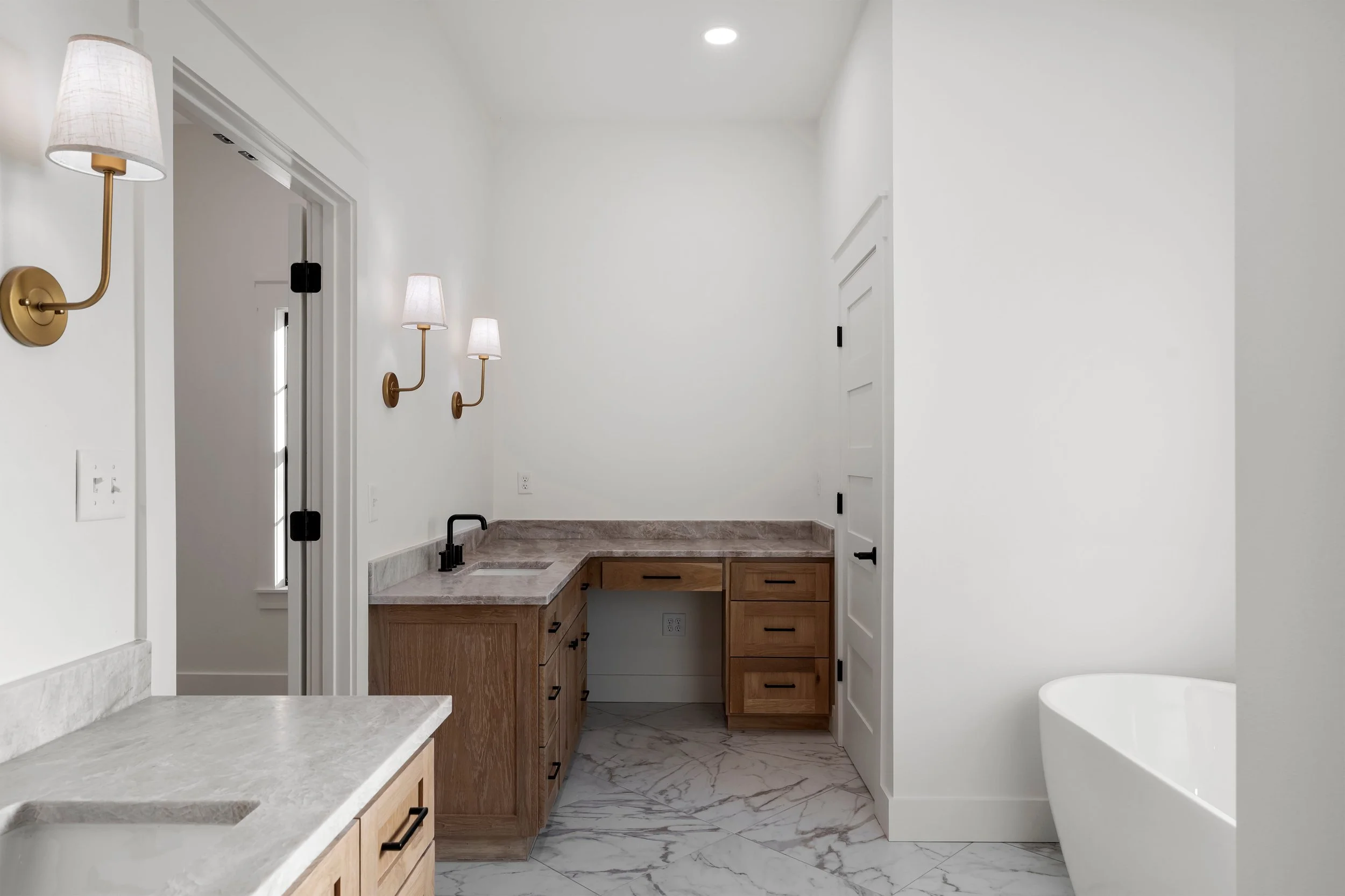 Bathroom with white walls, wooden cabinetry, marble flooring, and a white bathtub on the right. There are wall-mounted lamps on the left wall, a small sink area, and a white door.