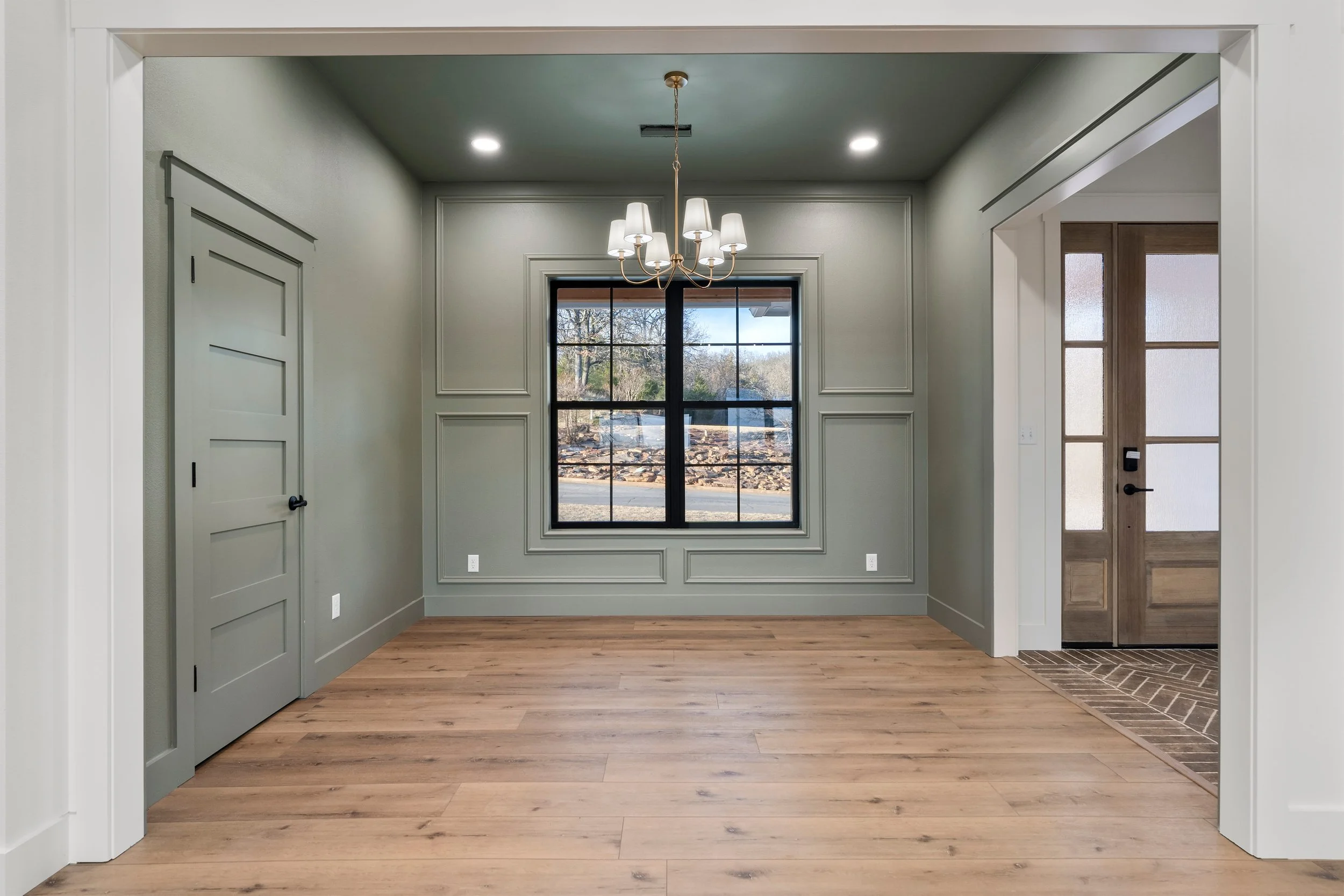 Empty living room with green walls, wooden floors, a window, a chandelier, and a door leading outside.