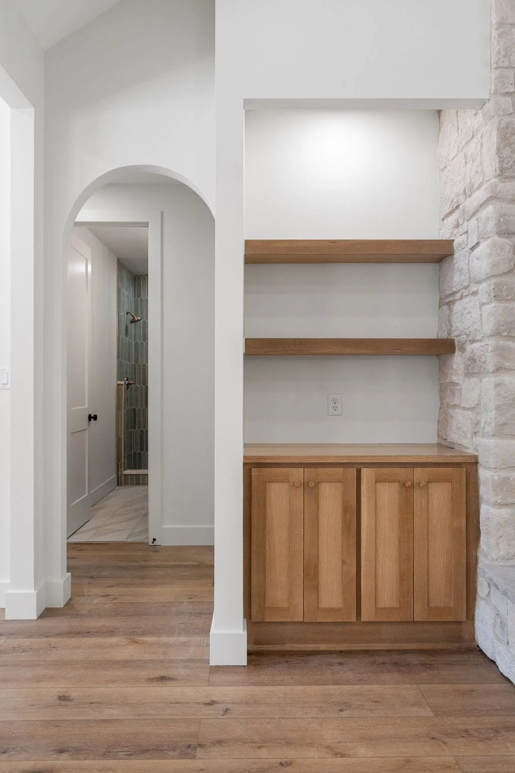 Interior of a home with natural wood flooring, a white wall with built-in wooden shelves, and a stone accent wall. A doorway leads to a bathroom with a shower featuring green patterned tiles.
